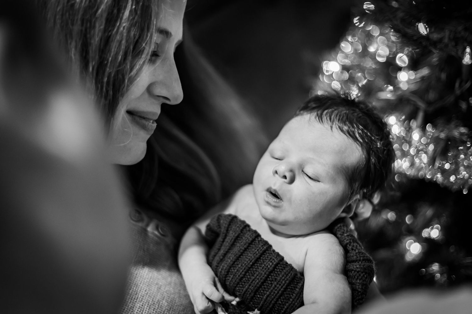 A tender black and white portrait of a sleeping newborn in a knit sweater with Christmas tree lights twinkling in background.