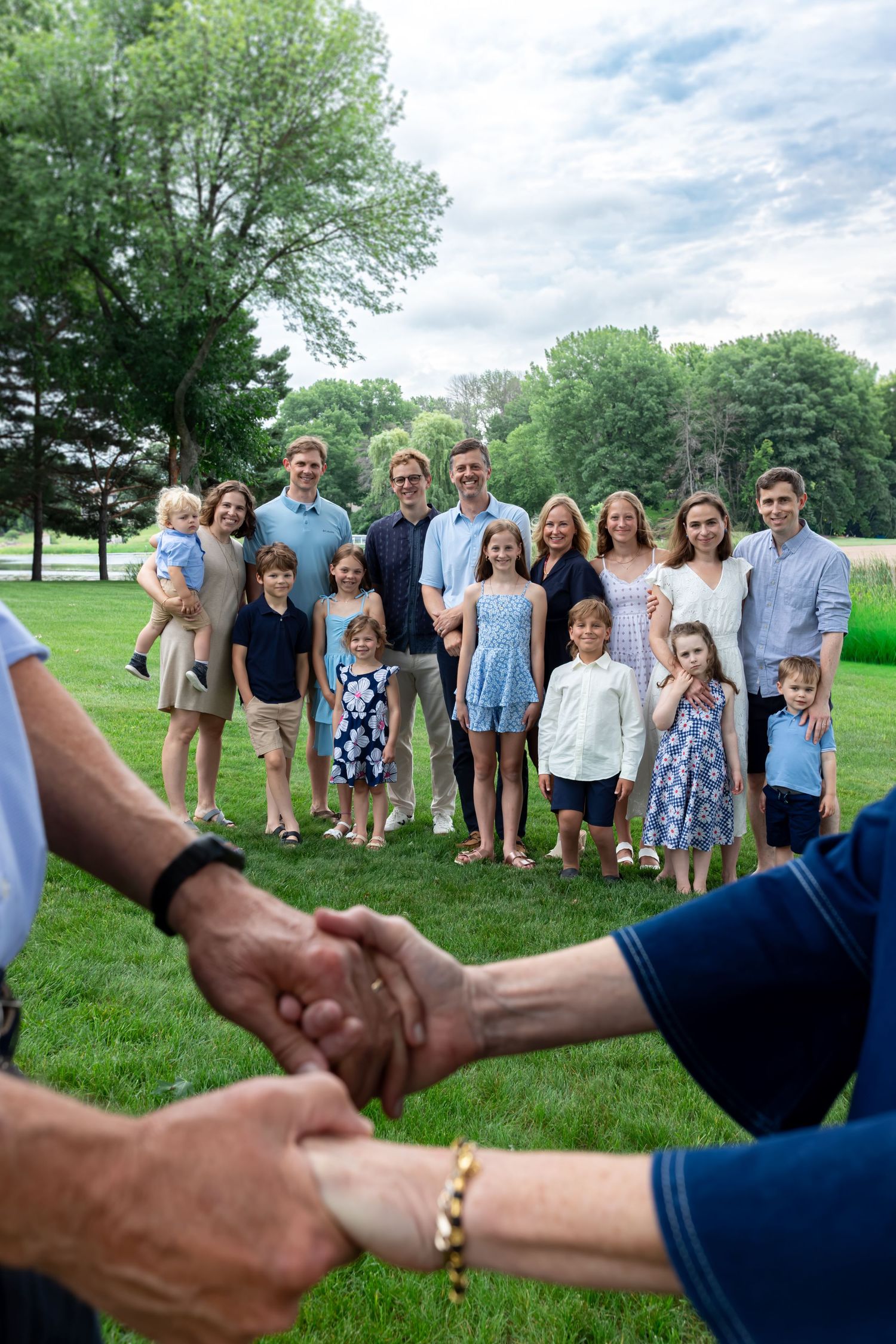 Elderly couple holds hands in foreground while large multigenerational family poses on green lawn with trees behind.
