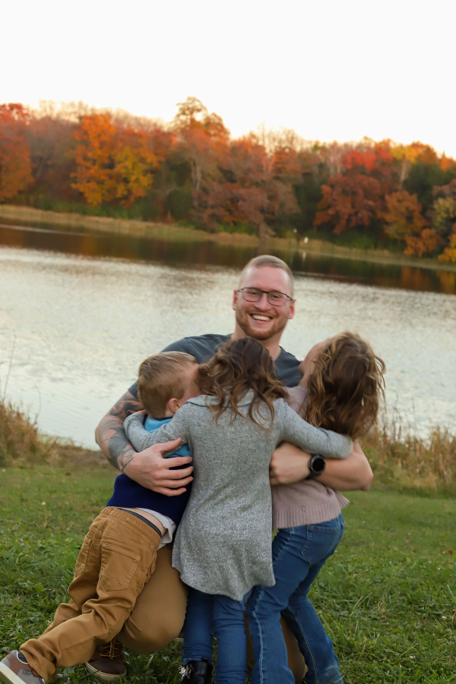 A joyful family shares hugs and laughter by an autumn lake with vibrant fall foliage in the background.