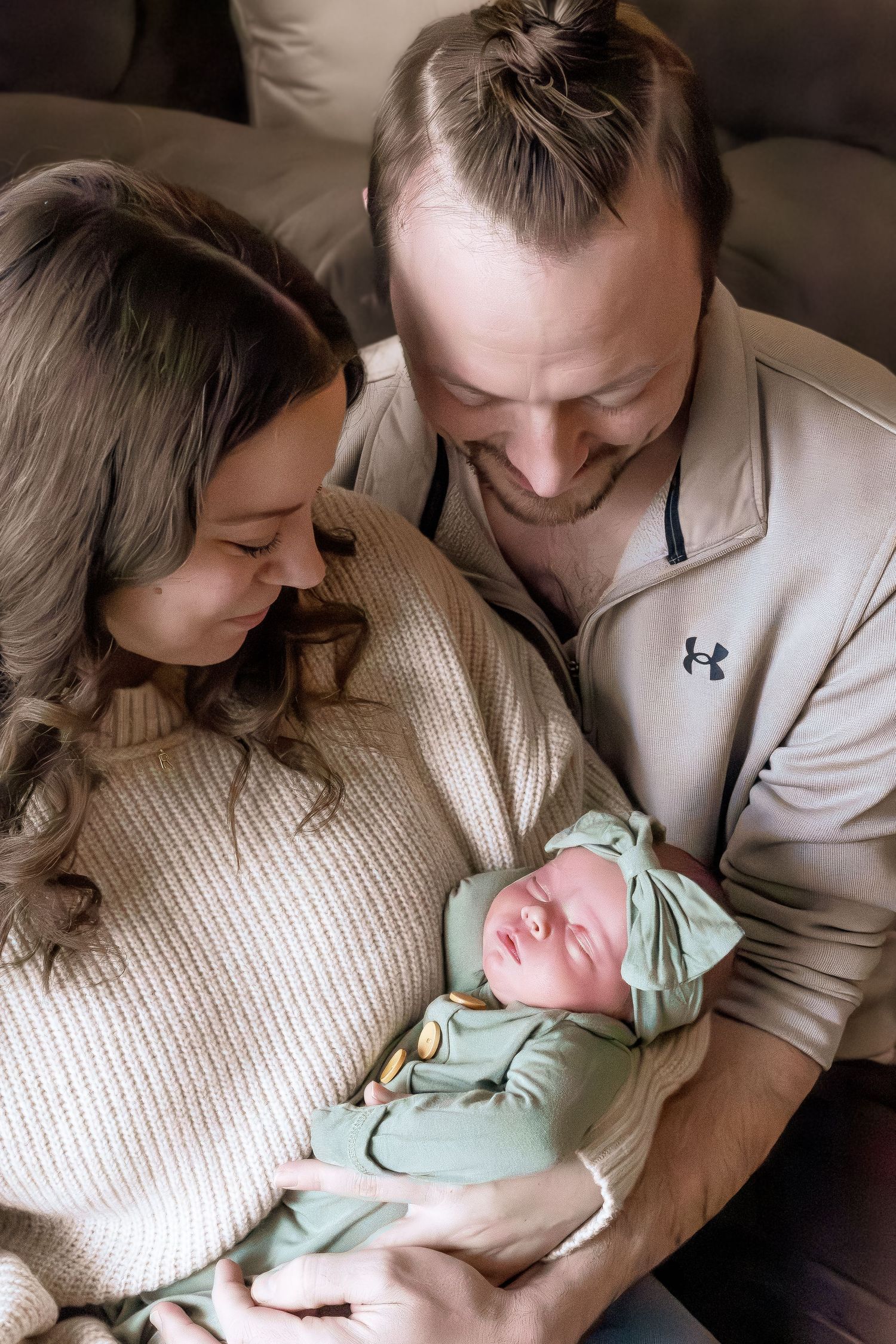 A couple in cozy sweaters lovingly gazes at their sleeping newborn baby wearing a mint green outfit.