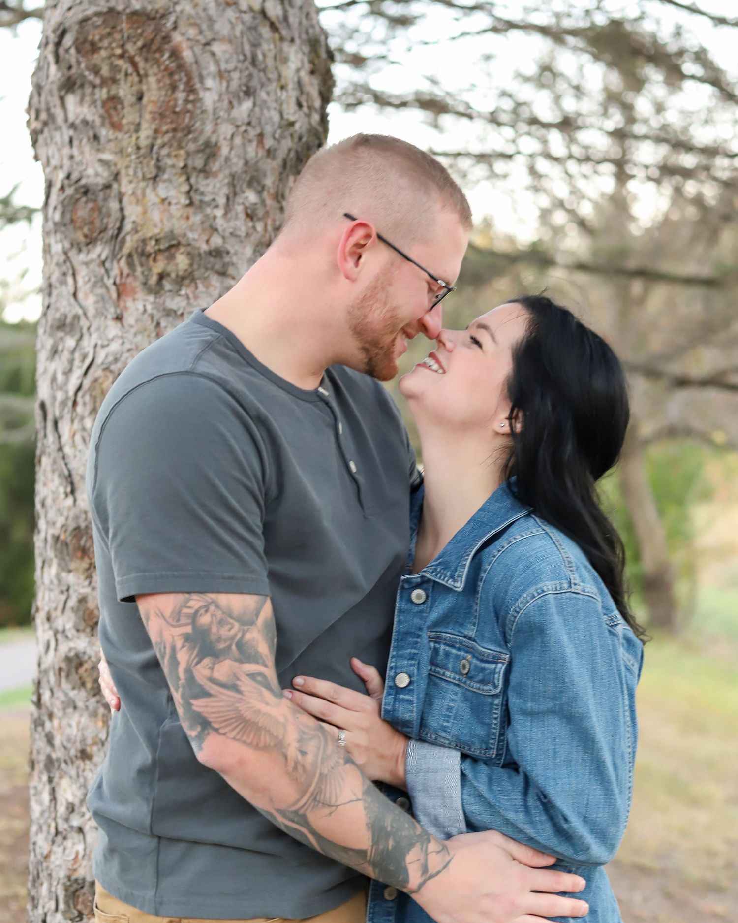 A couple in denim and grey clothing share intimate moments while posing against a tree trunk in an outdoor setting.