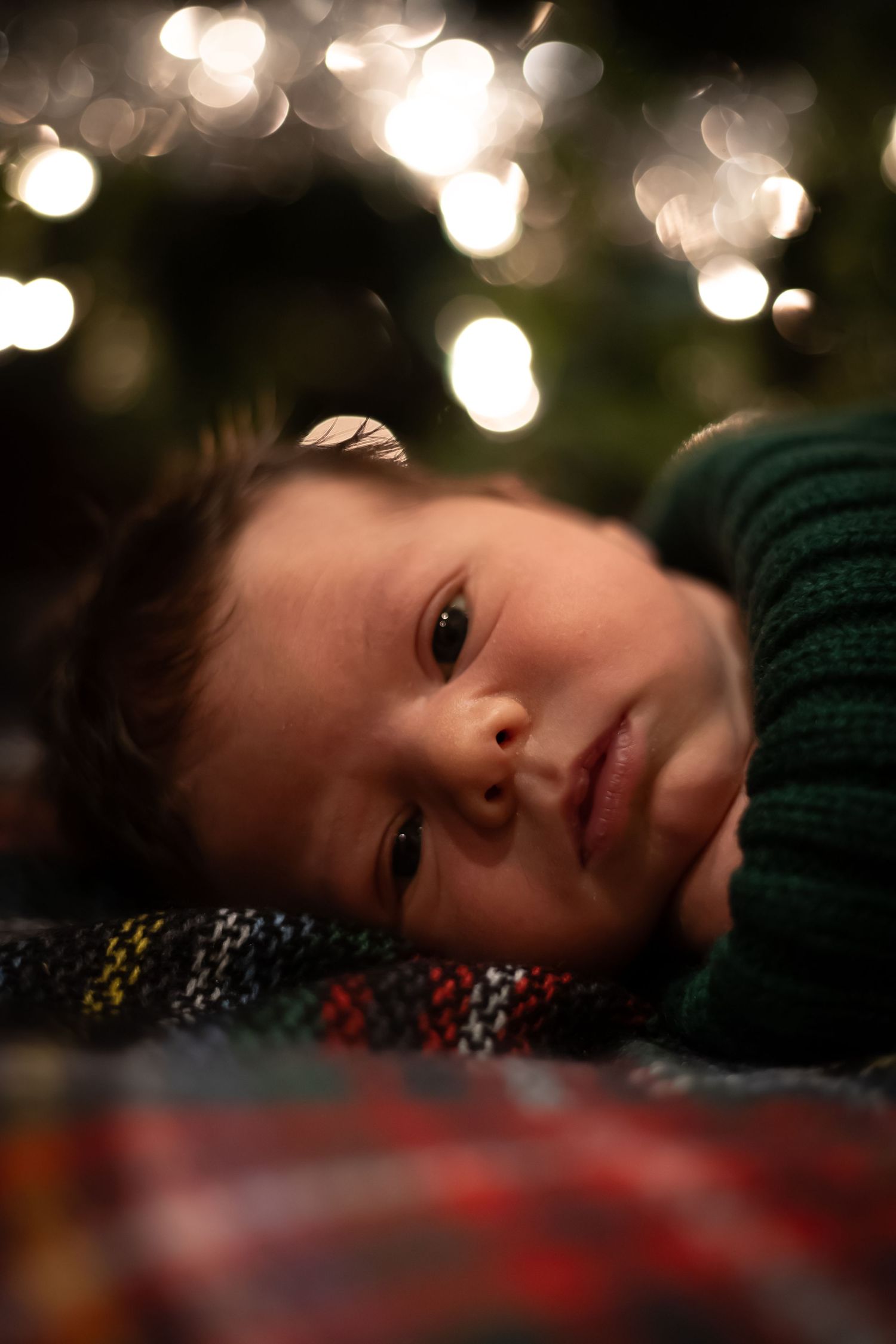 A child lies on a patterned surface with blurred holiday lights twinkling in the dark background.