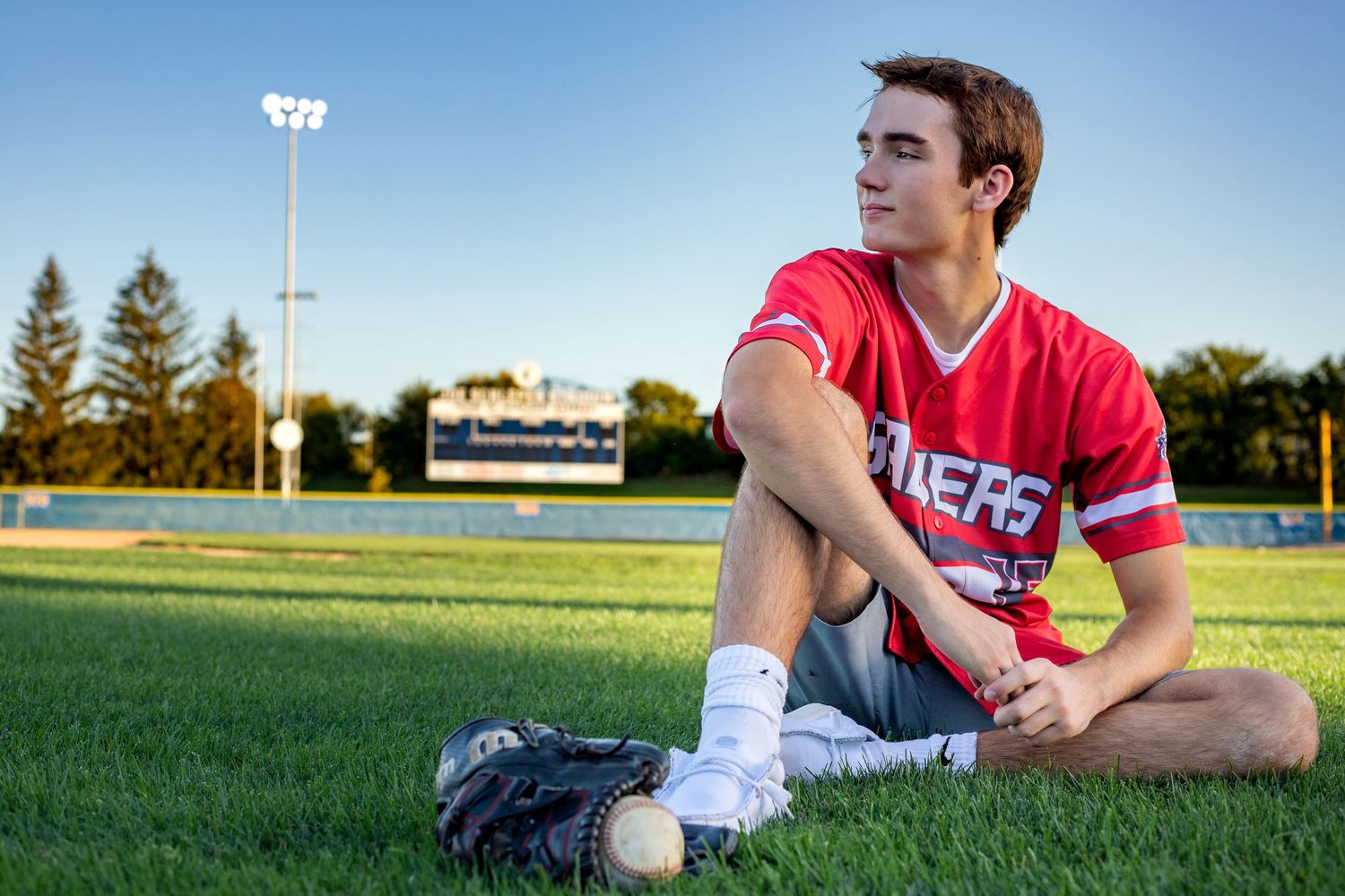 high-school-senior-photography-sitting-on-baseball-field-glove-and-ball-next-to-feet-lights-on-in-background.jpg