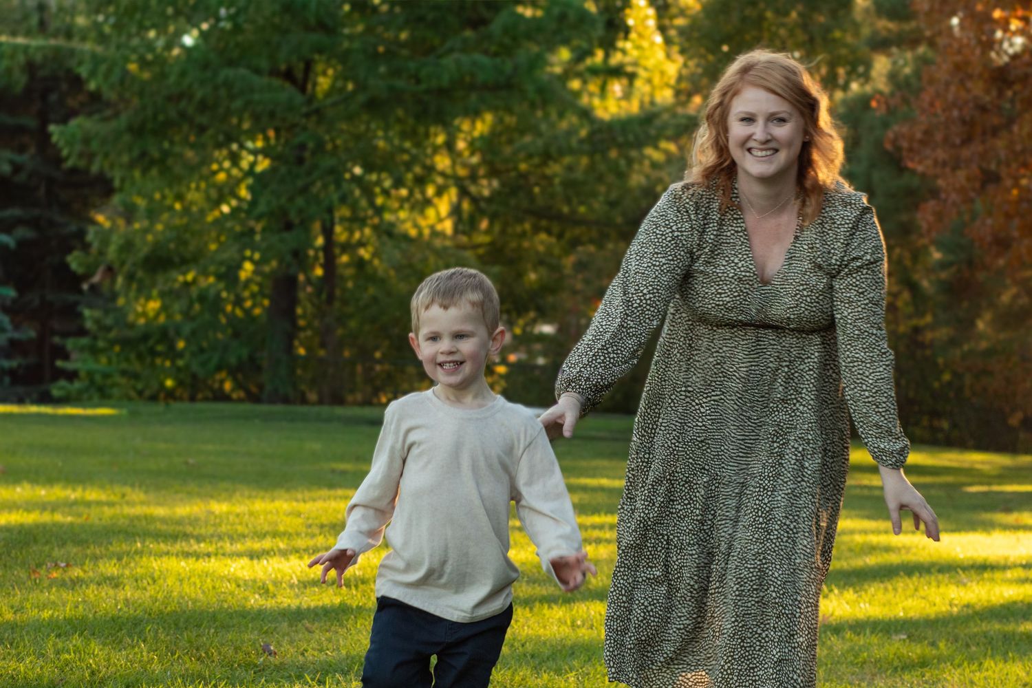A parent and child hold hands while walking together on a sunlit green lawn with autumn trees in the background.