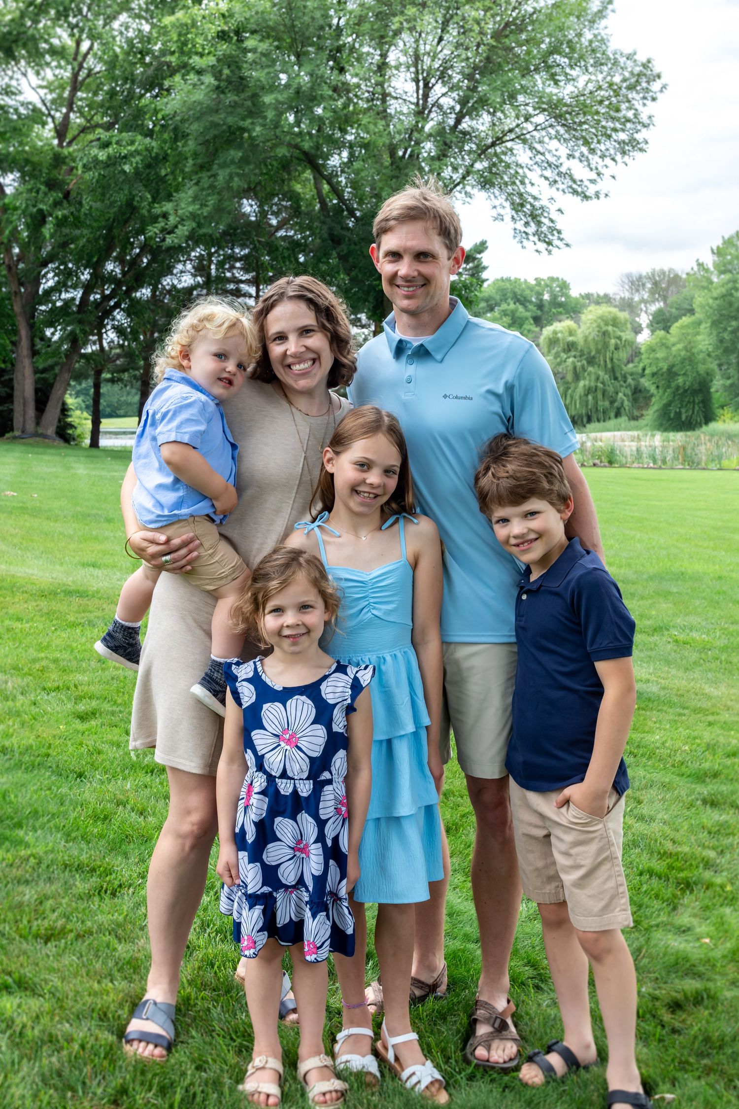 Happy family of six posing outdoors on green grass, parents with four young children, dressed in casual summer clothing.