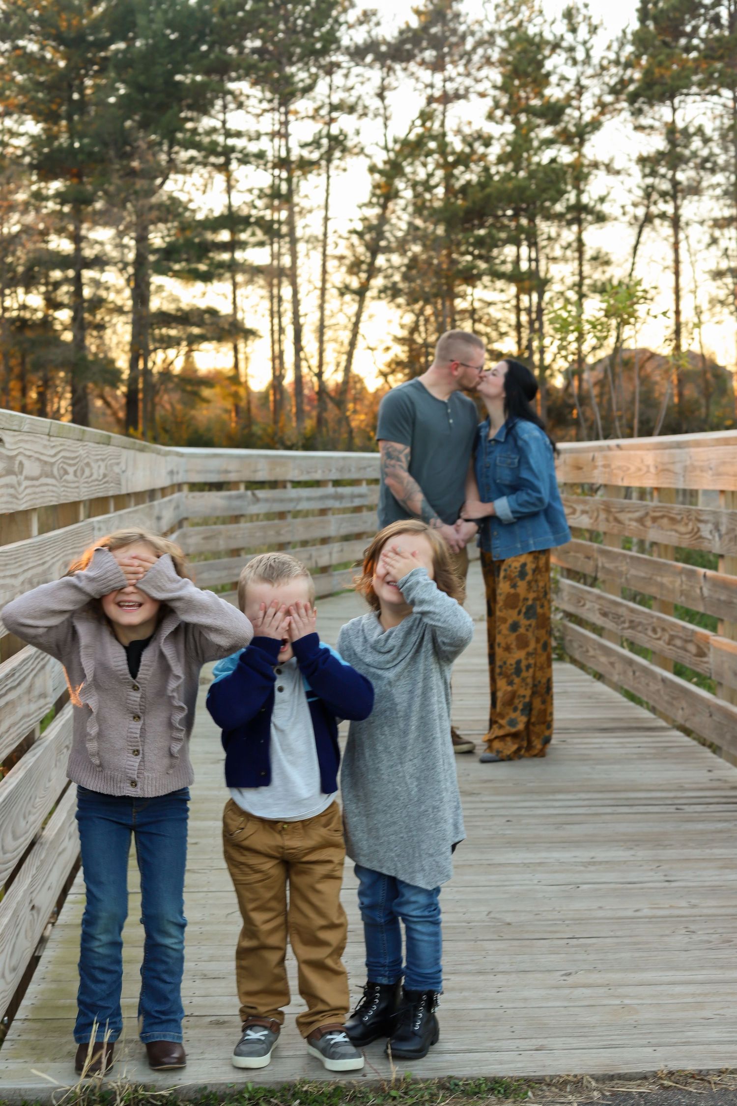 A playful family moment captured on a wooden boardwalk surrounded by tall pine trees at sunset.