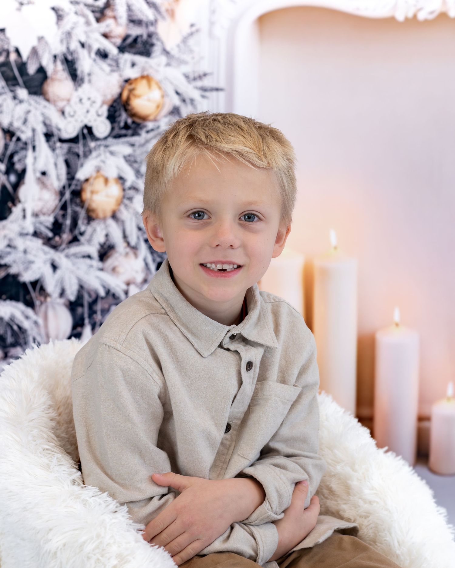 A young child in a beige collared shirt poses against a white winter holiday backdrop with lit candles and decorations.