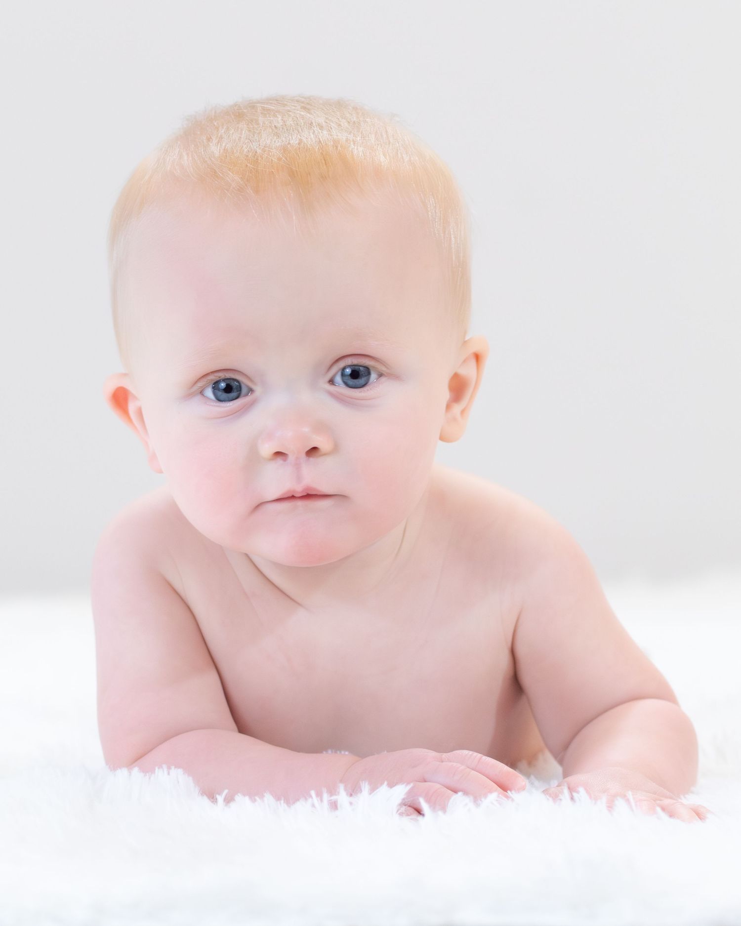 Baby with light hair and blue eyes lying on soft white blanket in studio setting.