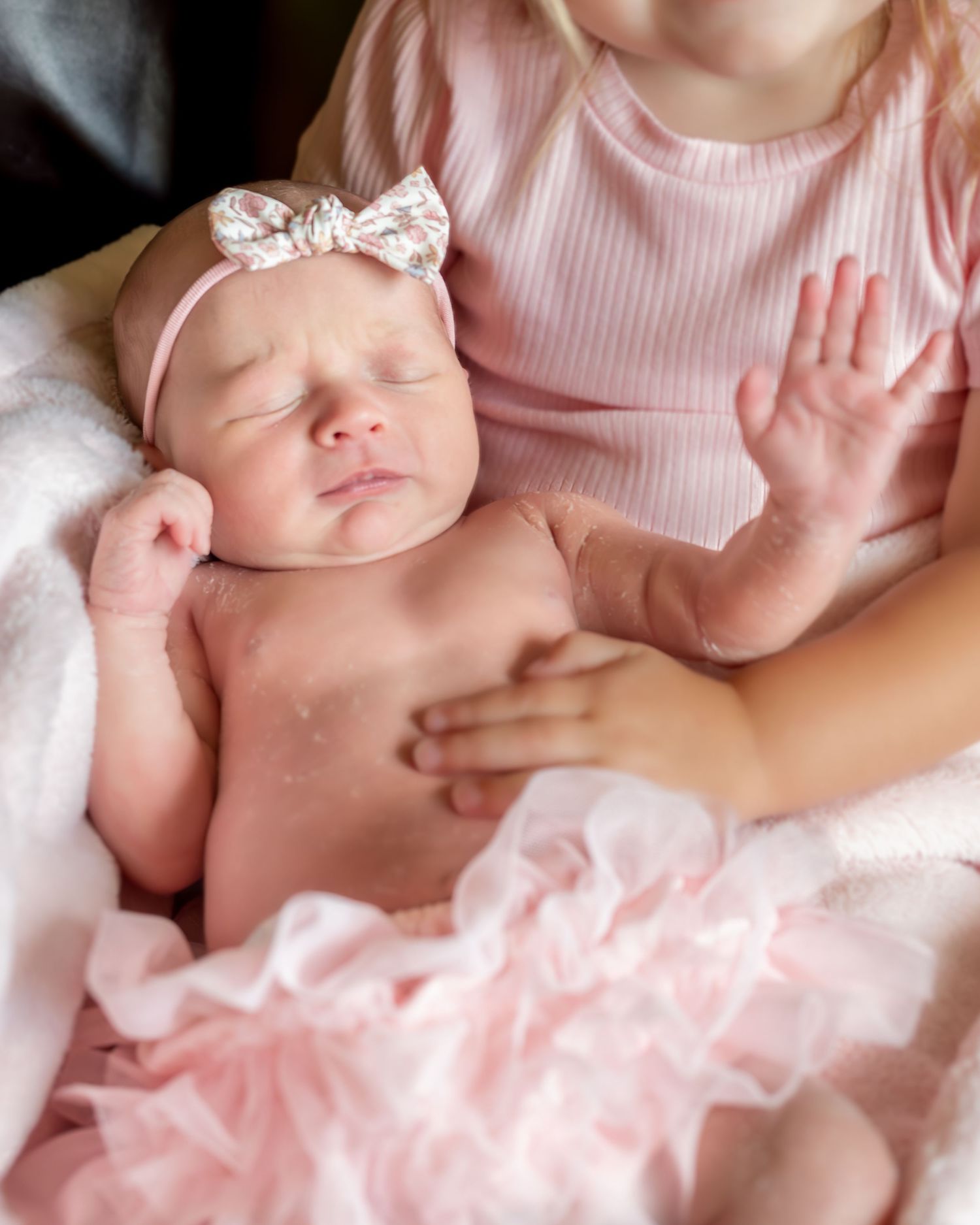 A sleeping newborn wearing a white bow headband rests on a pink blanket while being cradled in someone's arms.
