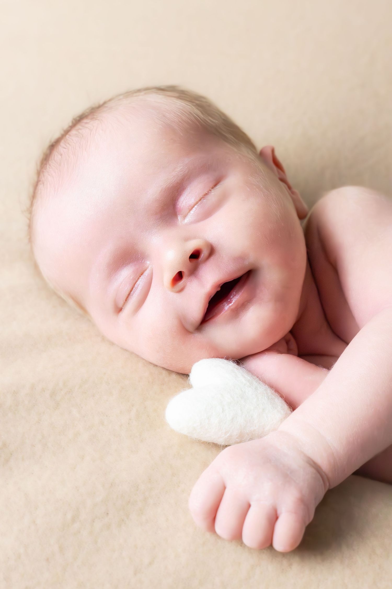 A newborn baby rests peacefully on cream-colored fabric in gentle lighting.