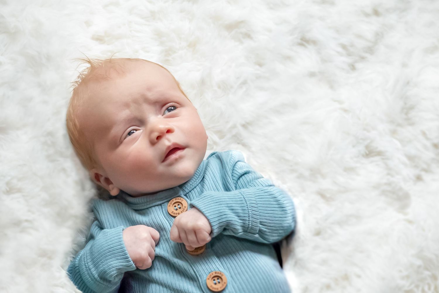 A newborn baby wearing a blue sweater lies on a soft white blanket while looking upward.