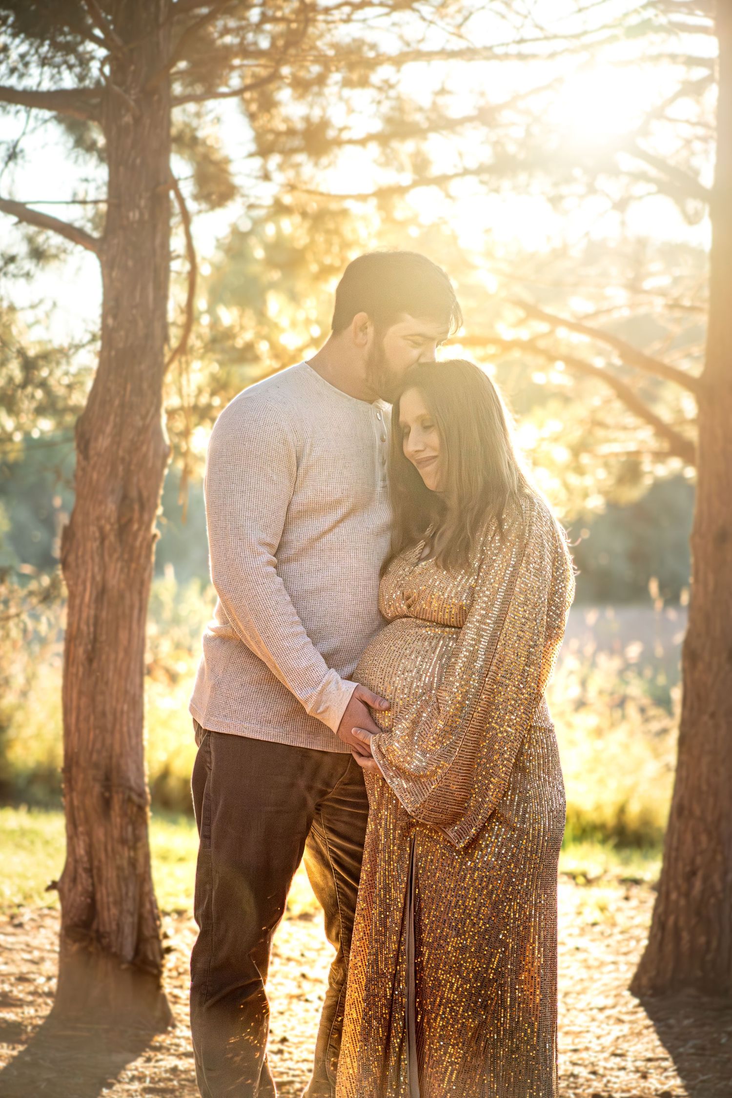 Romantic maternity photo of couple in warm sunset light among pine trees.