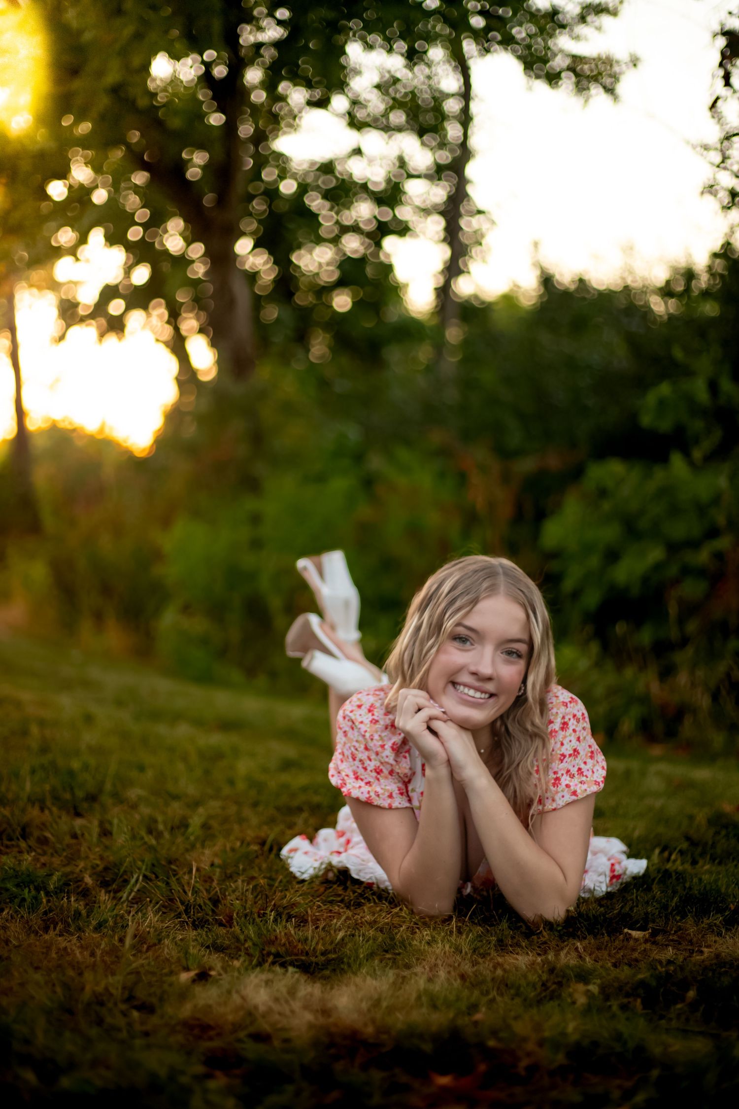 Young blonde woman in floral dress lying on grass smiling at sunset in a lush green park.