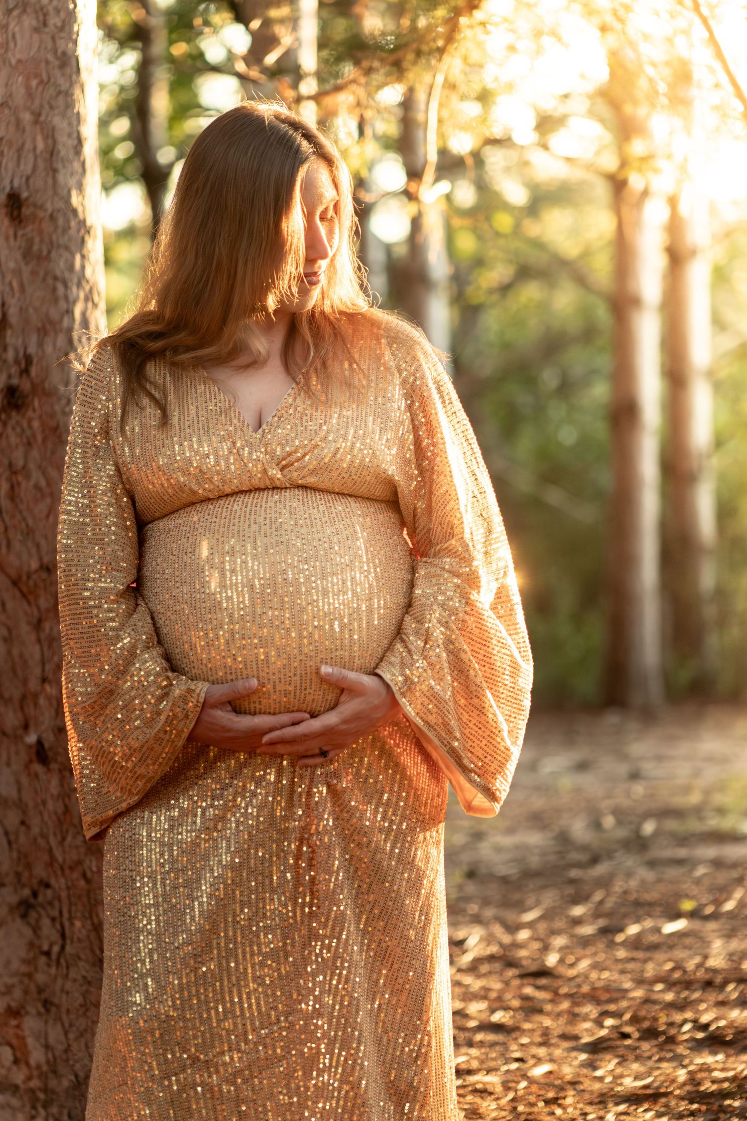 Side view of pregnant woman in shimmering gold dress standing in forest sunlight.