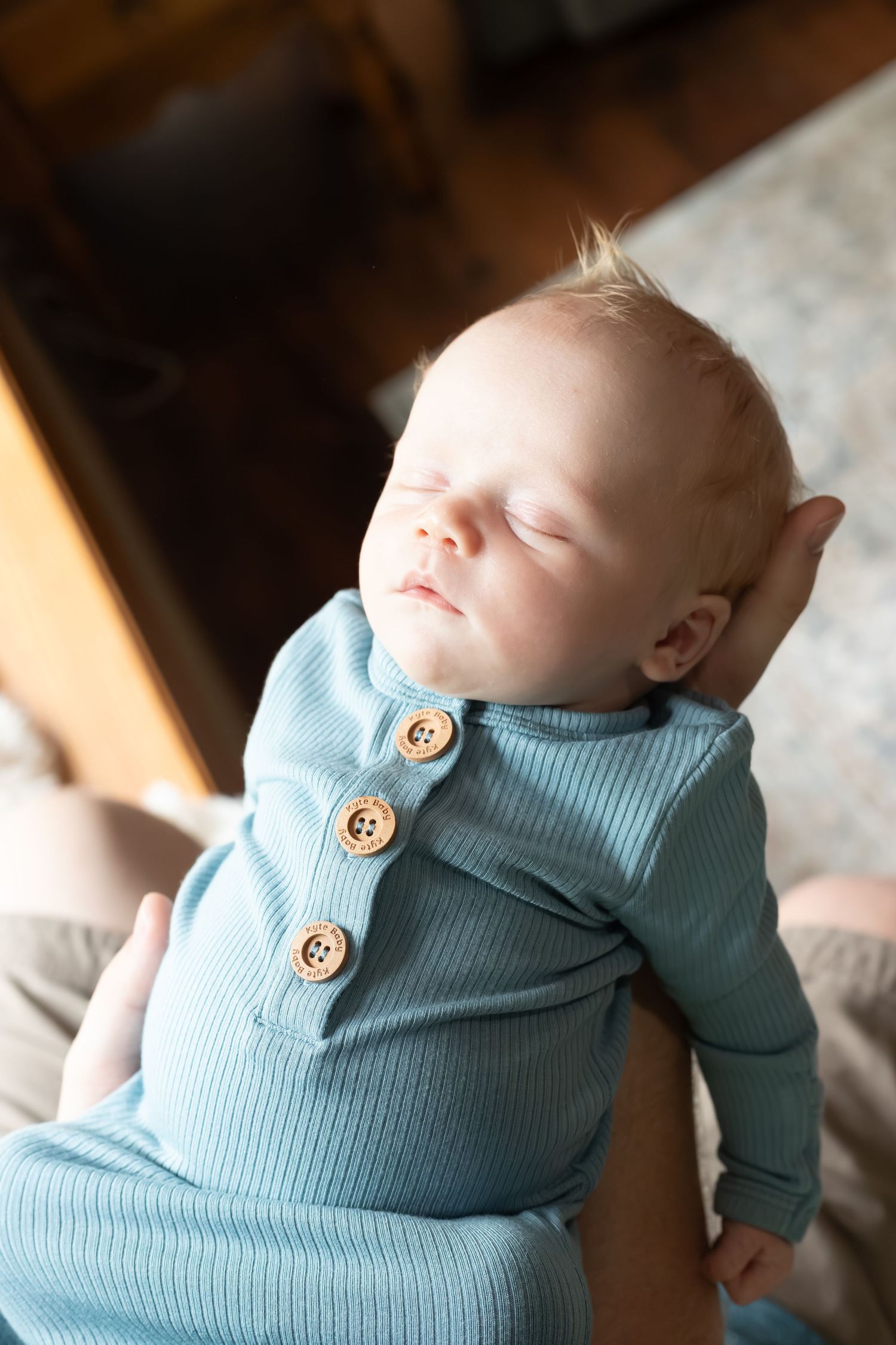 Sleeping baby wearing cozy light blue ribbed outfit with wooden buttons rests peacefully in warm sunlight.