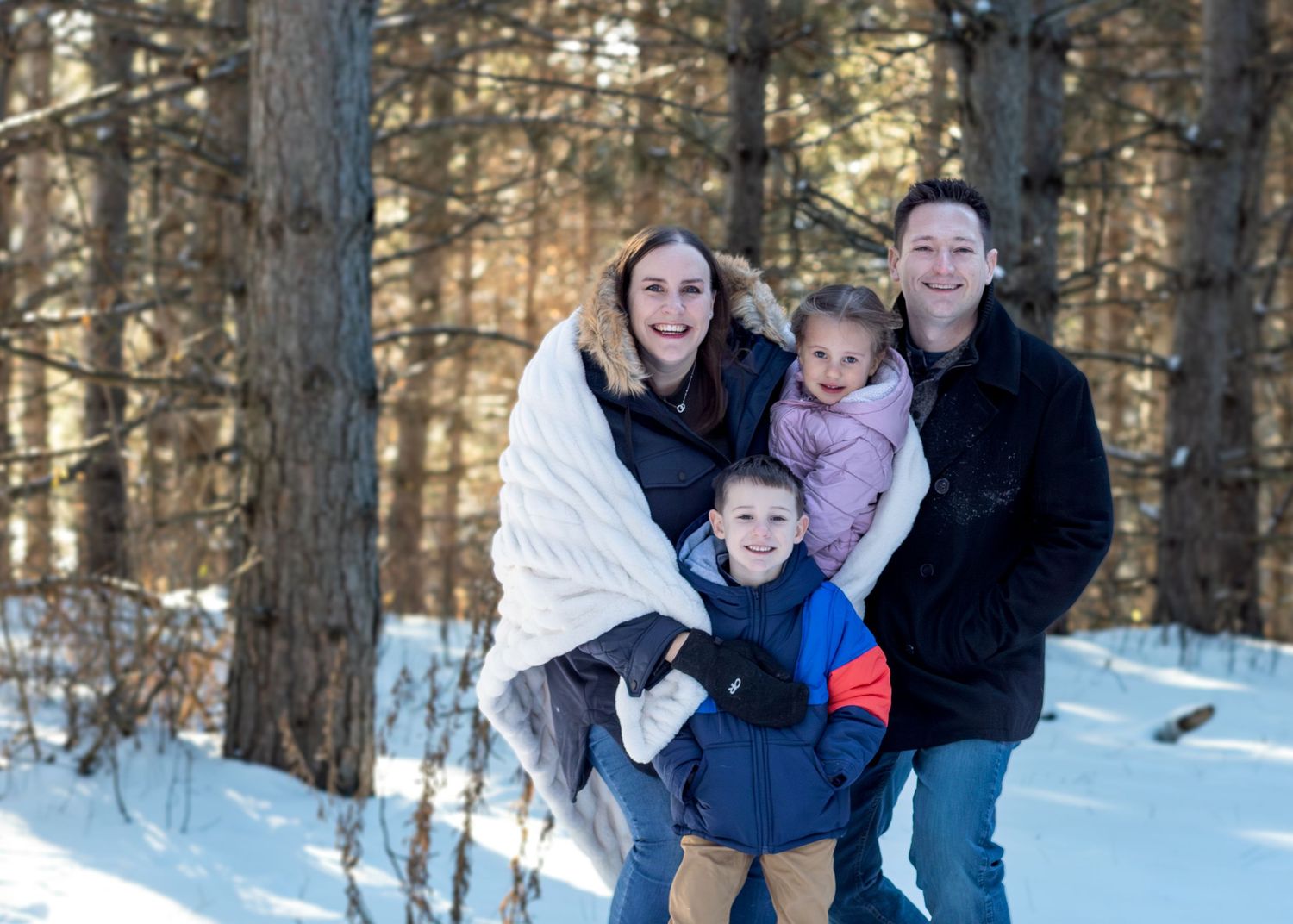 A family enjoys a winter day together in a snowy forest setting with pine trees in the background.