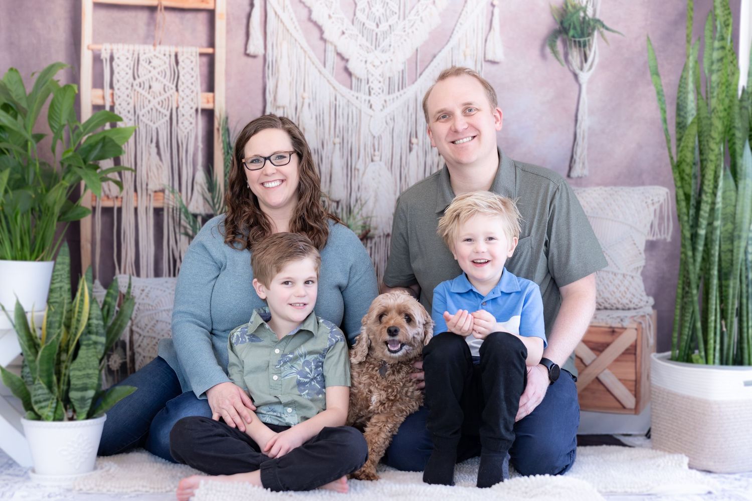 A family sits together on a couch with their brown dog, surrounded by indoor plants and macrame wall decor.
