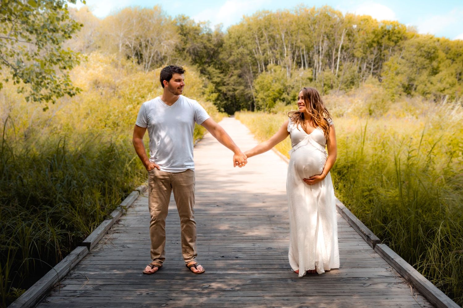 maternity photo, white dress, white shirt, holding hands across a boardwalk surrounded by greenery