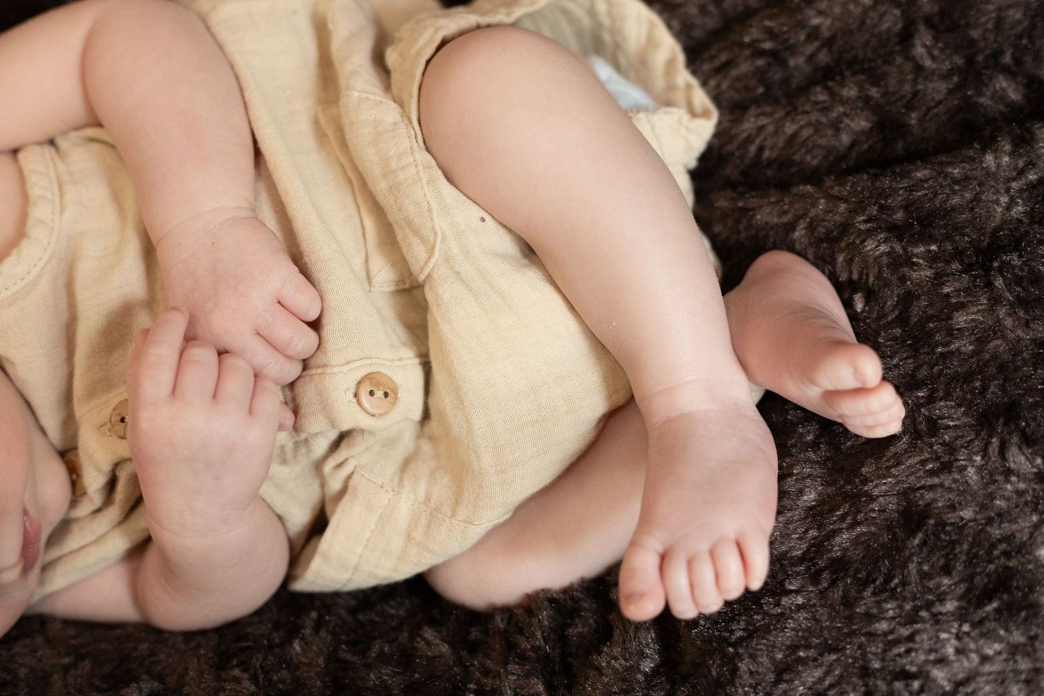 Close up of tiny baby feet and legs wearing a light beige outfit while resting on dark brown textured fabric.