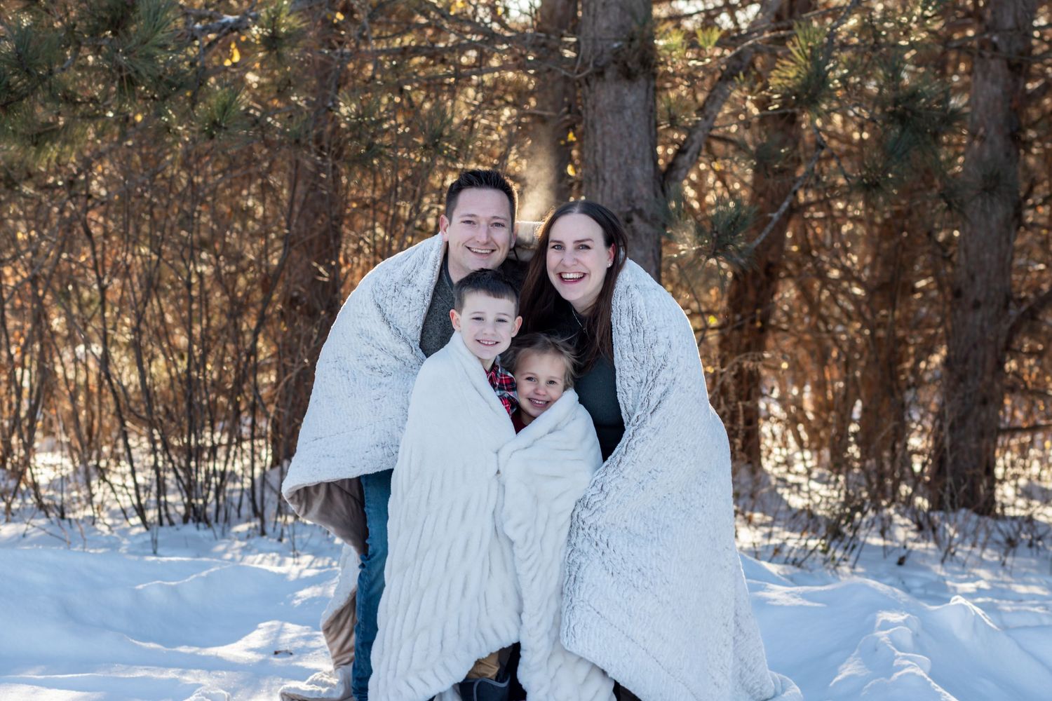 A couple wearing white winter coats embrace and laugh together in a snowy forest setting during a winter photoshoot.