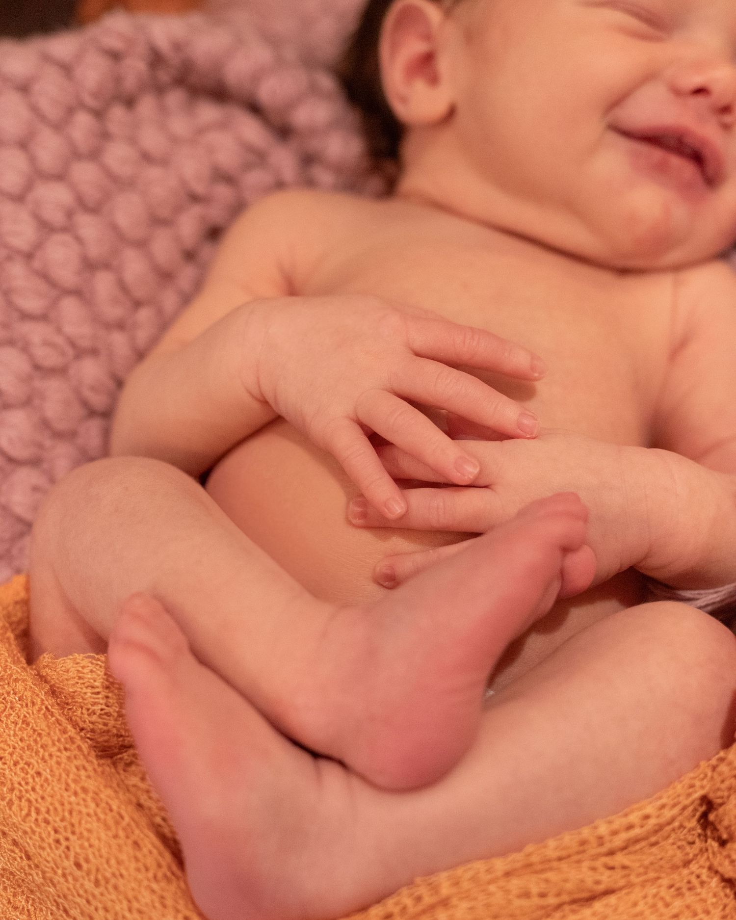 Sleeping infant rests on pink textured blanket with bare feet and hands visible against orange fabric background.