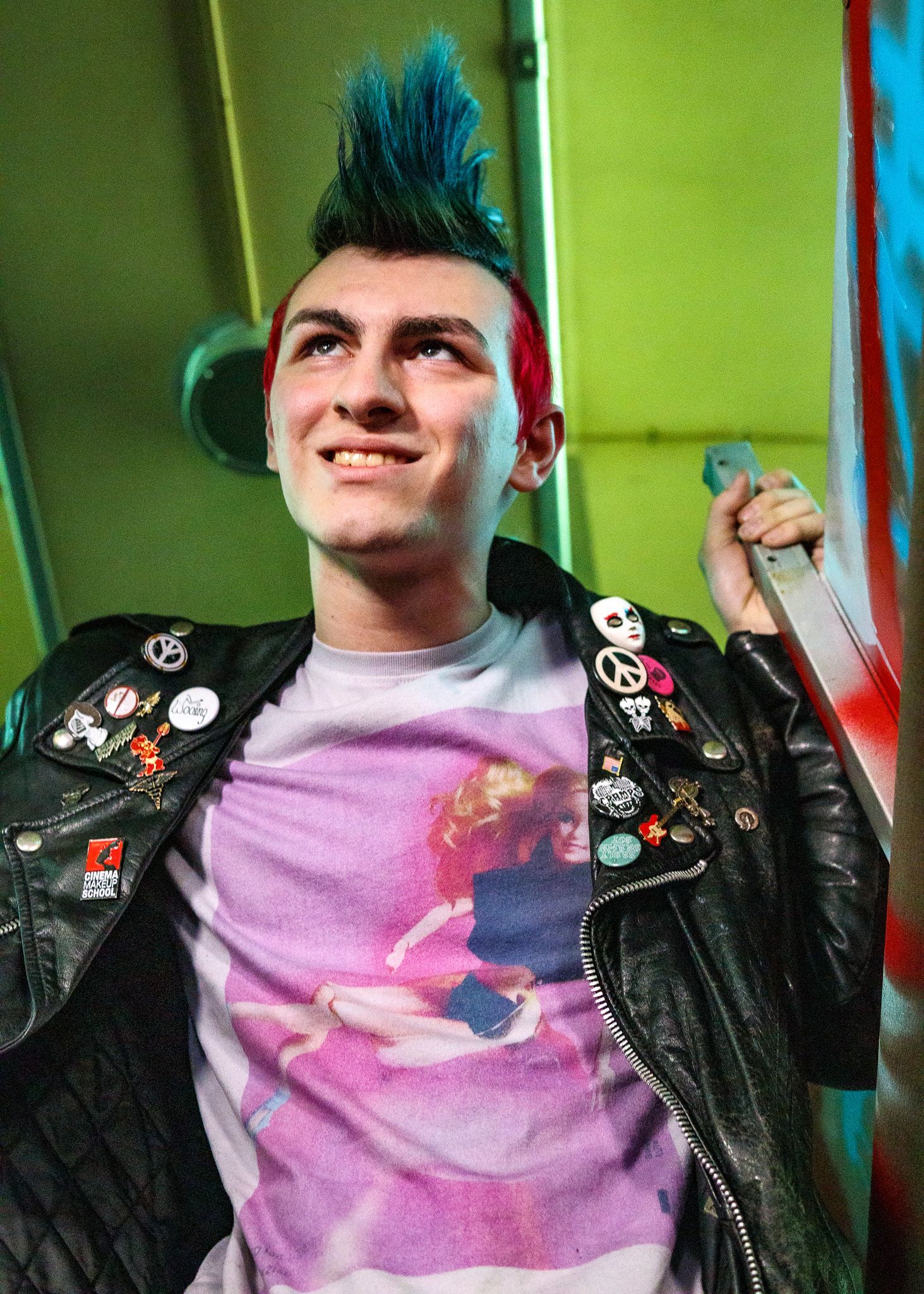Alternative style portrait with blue hair, leather jacket and pins against bright green wall.