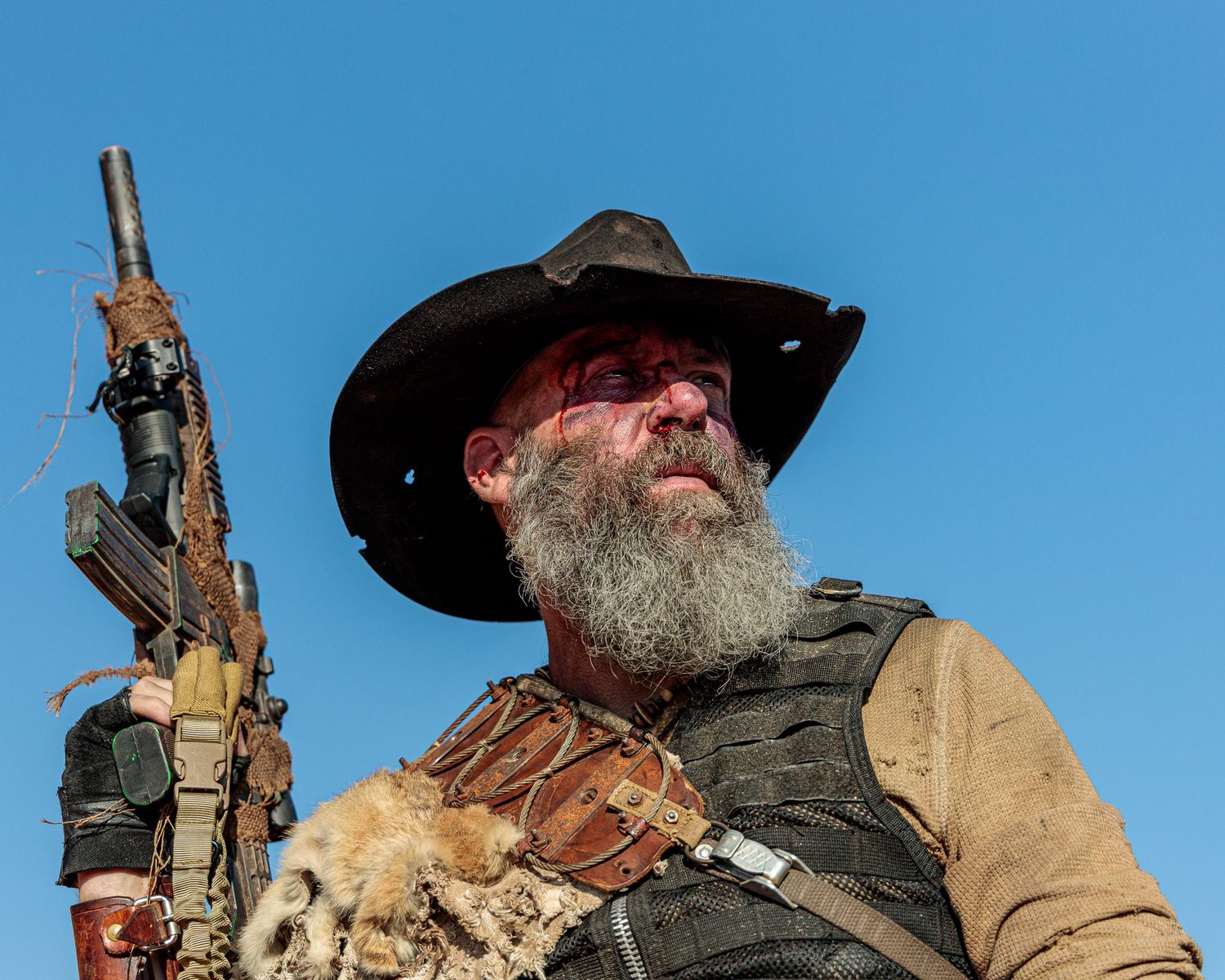 A rugged figure in tactical gear and cowboy hat poses against bright blue sky with weapon.