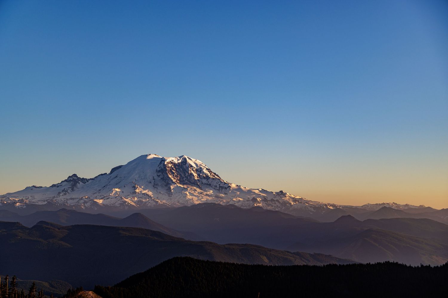 Mountain range silhouettes against blue sky with snow-capped peaks at dusk.