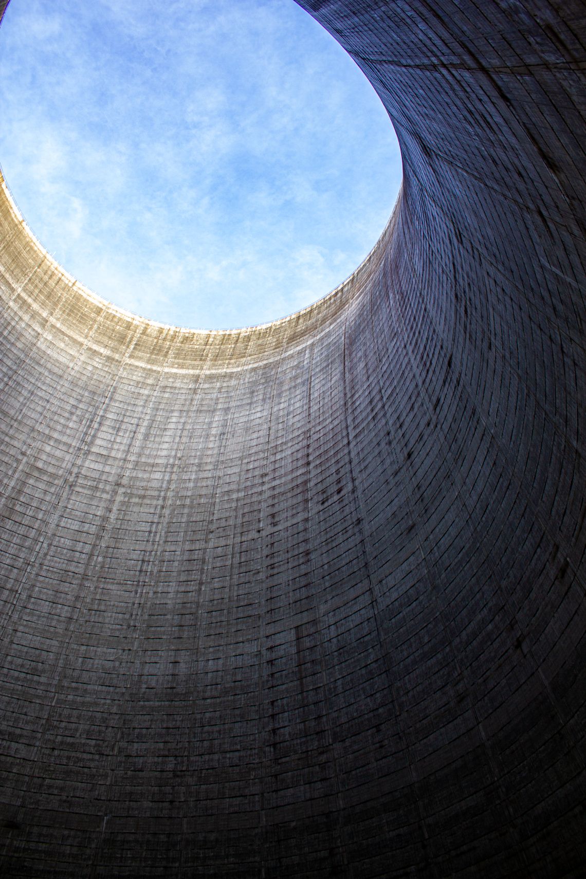 Looking up through a massive cooling tower reveals a circular opening against a bright blue sky.