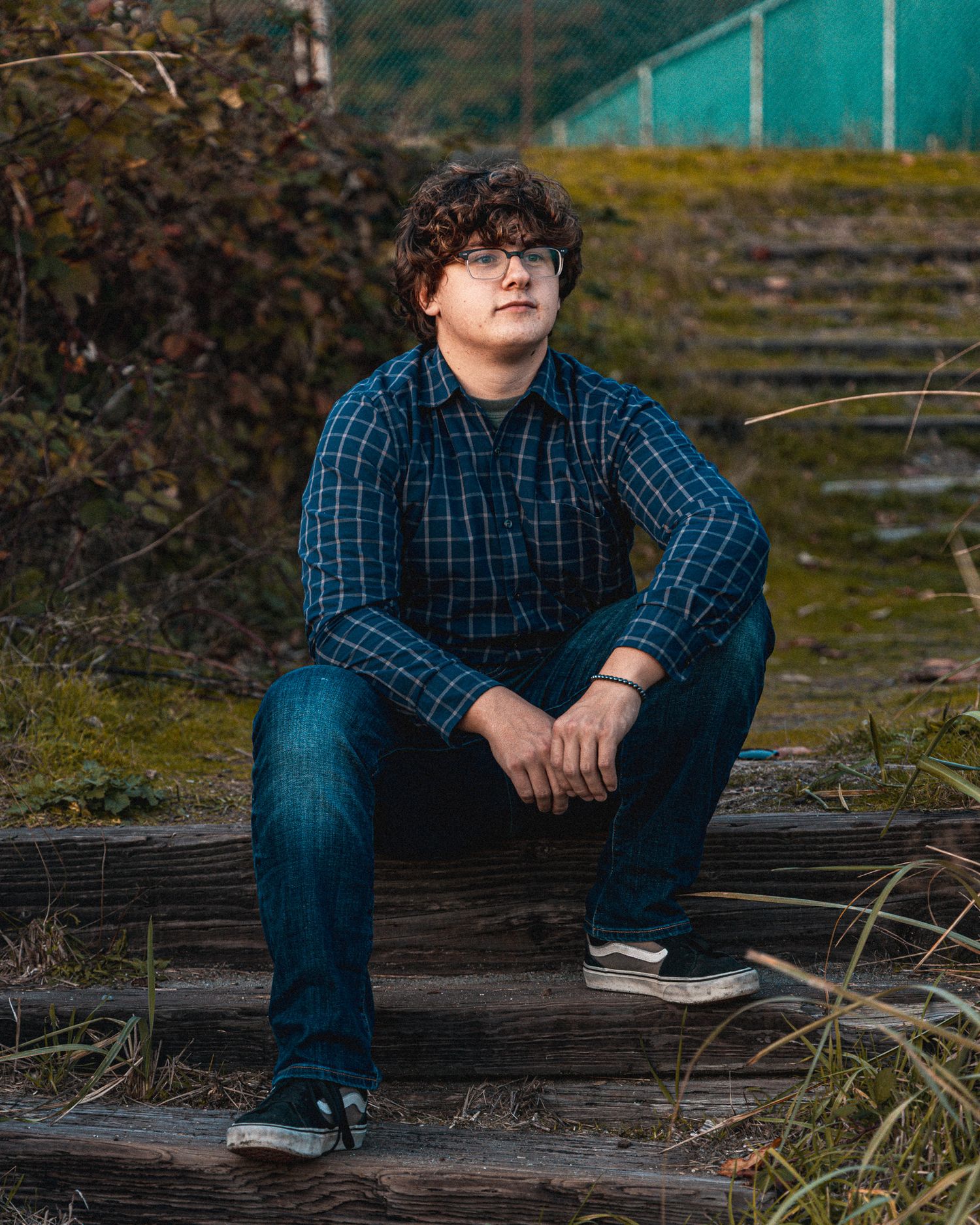 Person in blue plaid shirt and jeans sitting on wooden surface in outdoor setting.