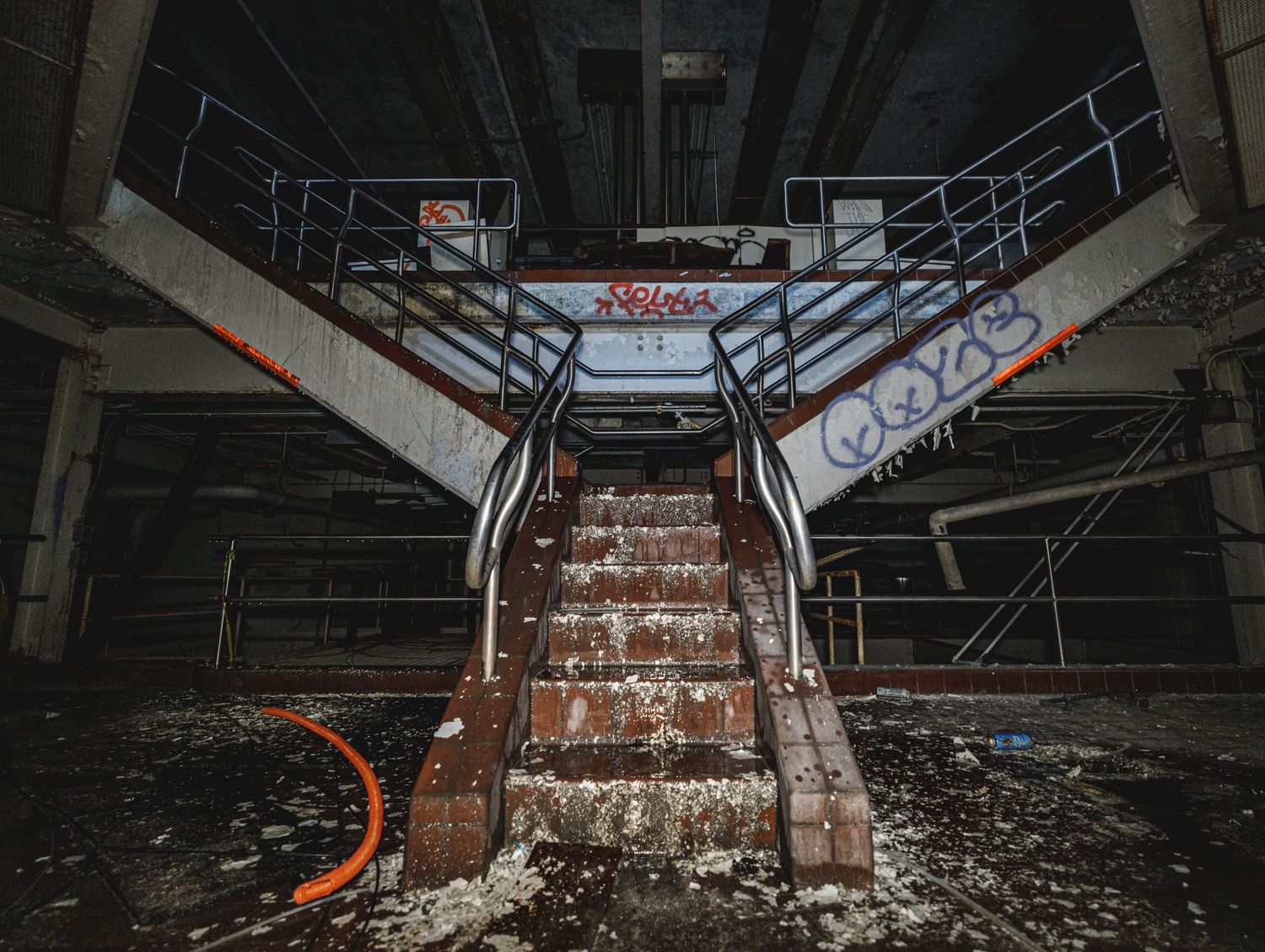 Dark industrial basement corridor with concrete stairs and exposed pipes creating symmetrical perspective.