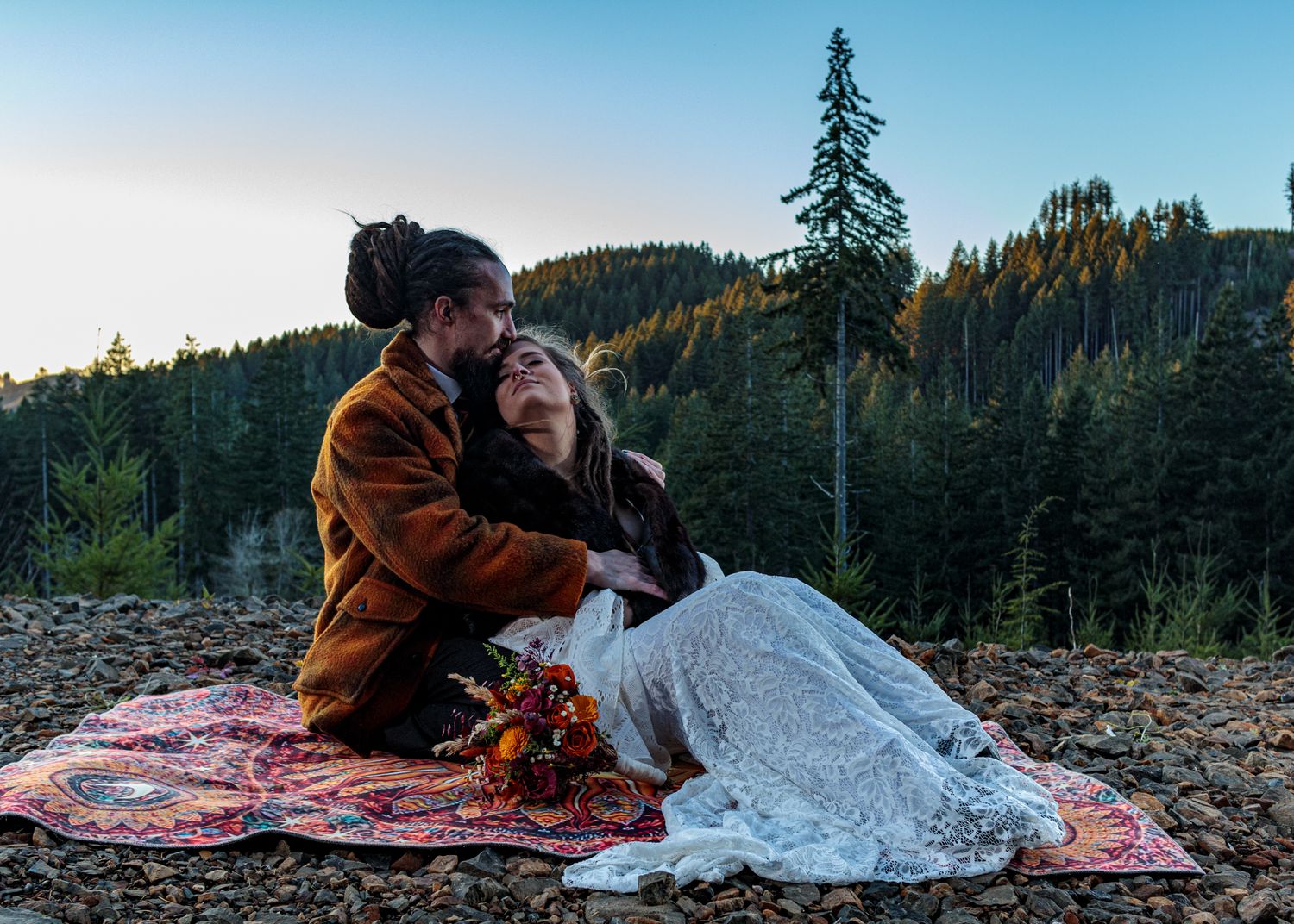 A couple poses on a blanket during sunset with evergreen trees and mountains in view.