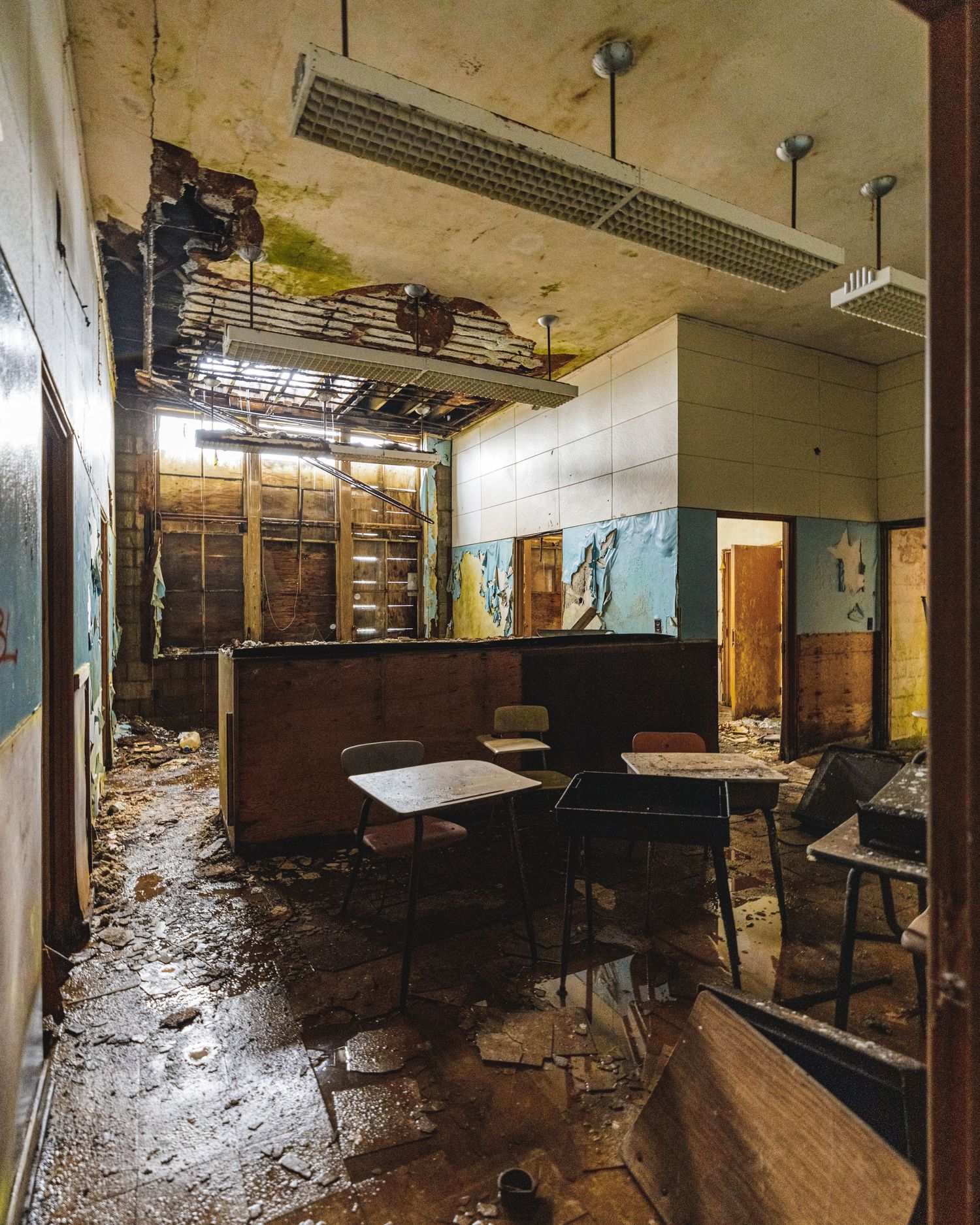 Abandoned school cafeteria with deteriorating ceiling, debris-covered floor, overturned chairs, and decaying walls in a state of disrepair.