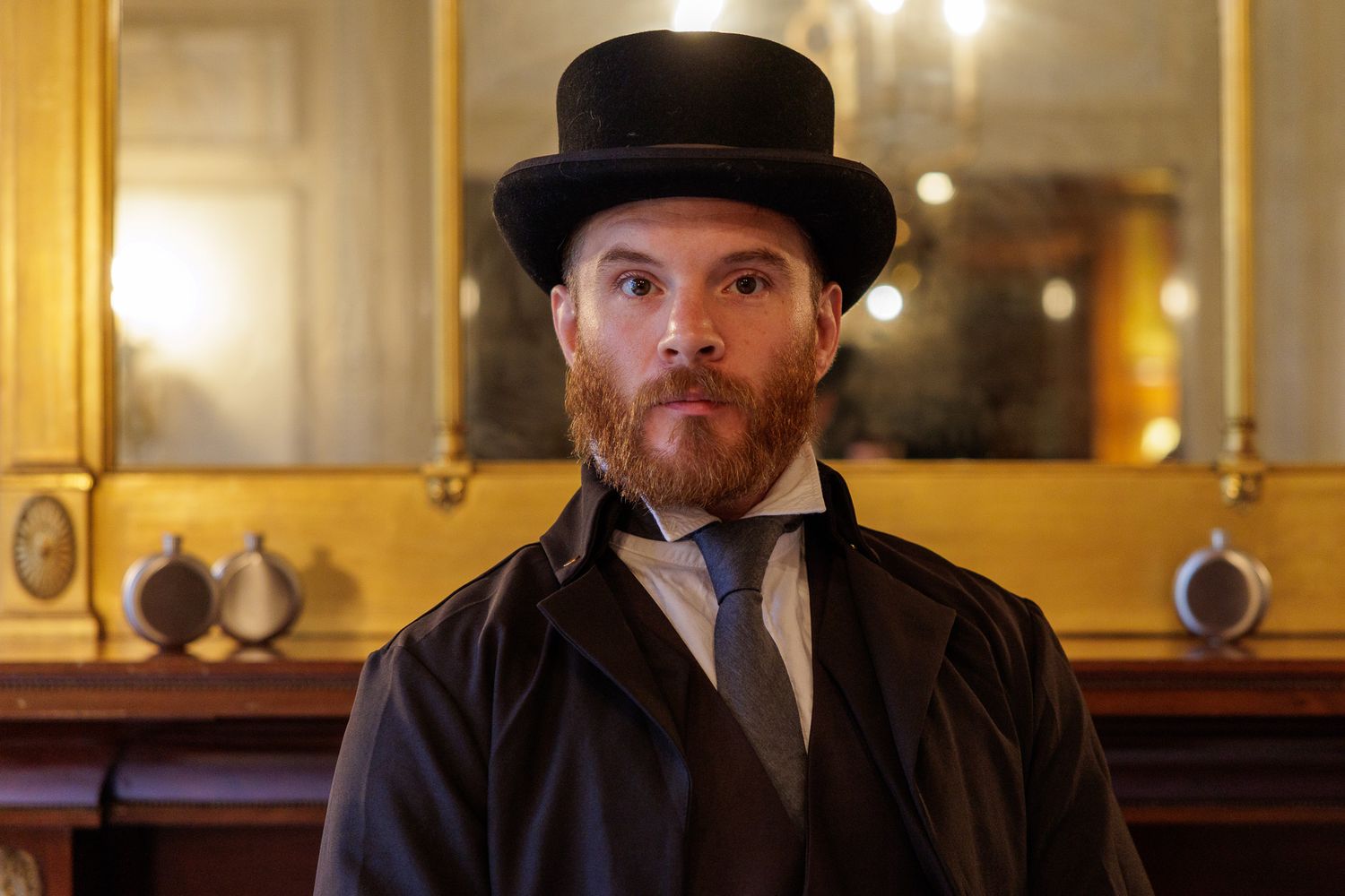 A person in Victorian attire with a top hat and beard poses in an elegant indoor setting.