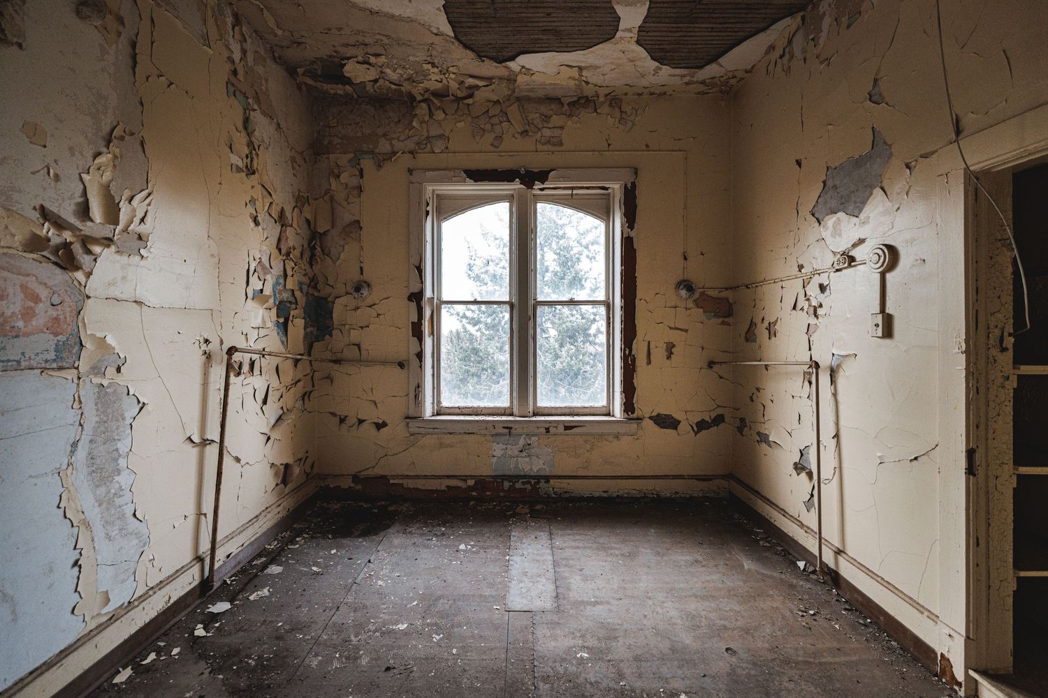 Abandoned hospital corridor with peeling walls and debris scattered across the floor in natural light.