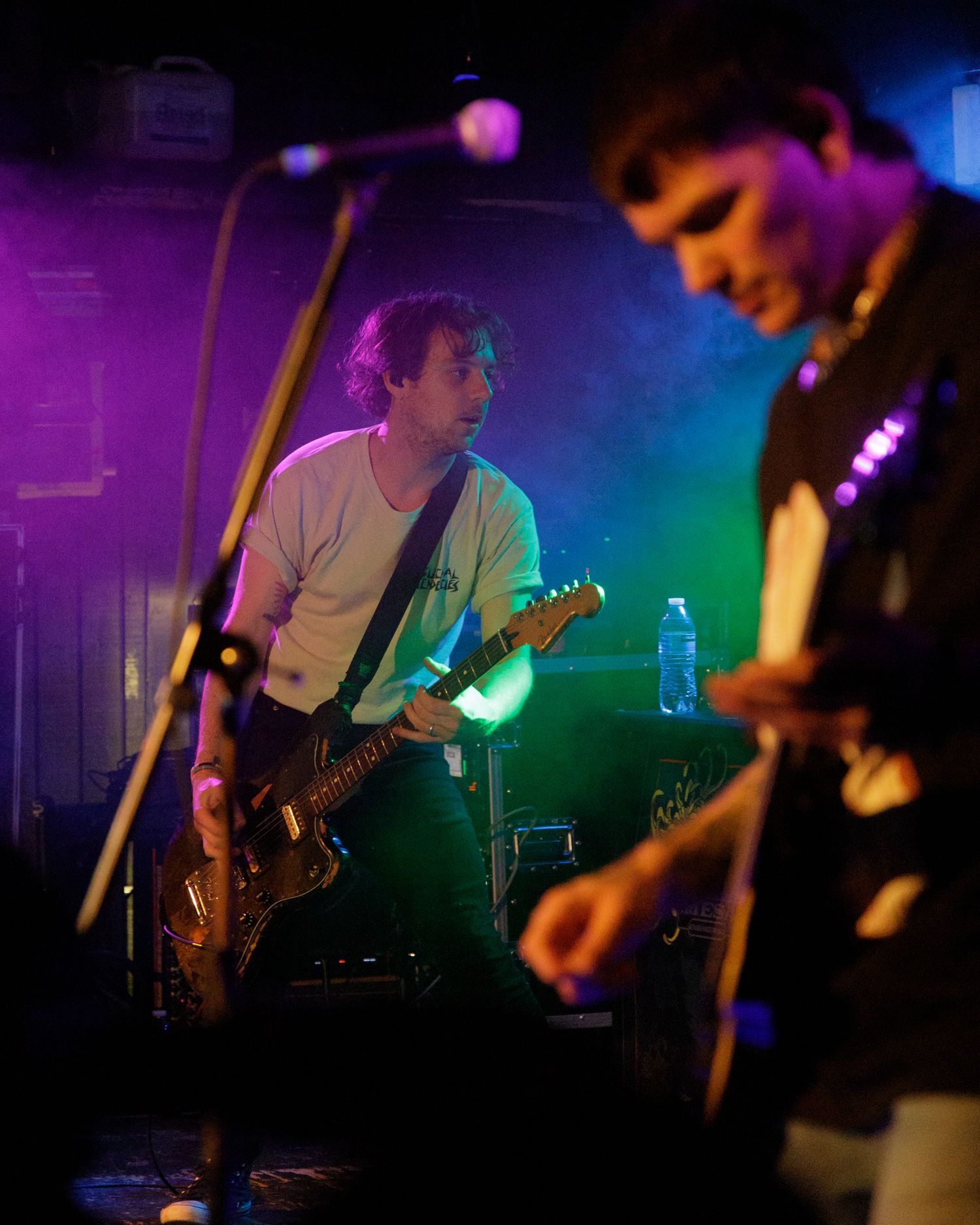Musicians perform on dimly lit stage with colorful purple and green lighting effects.