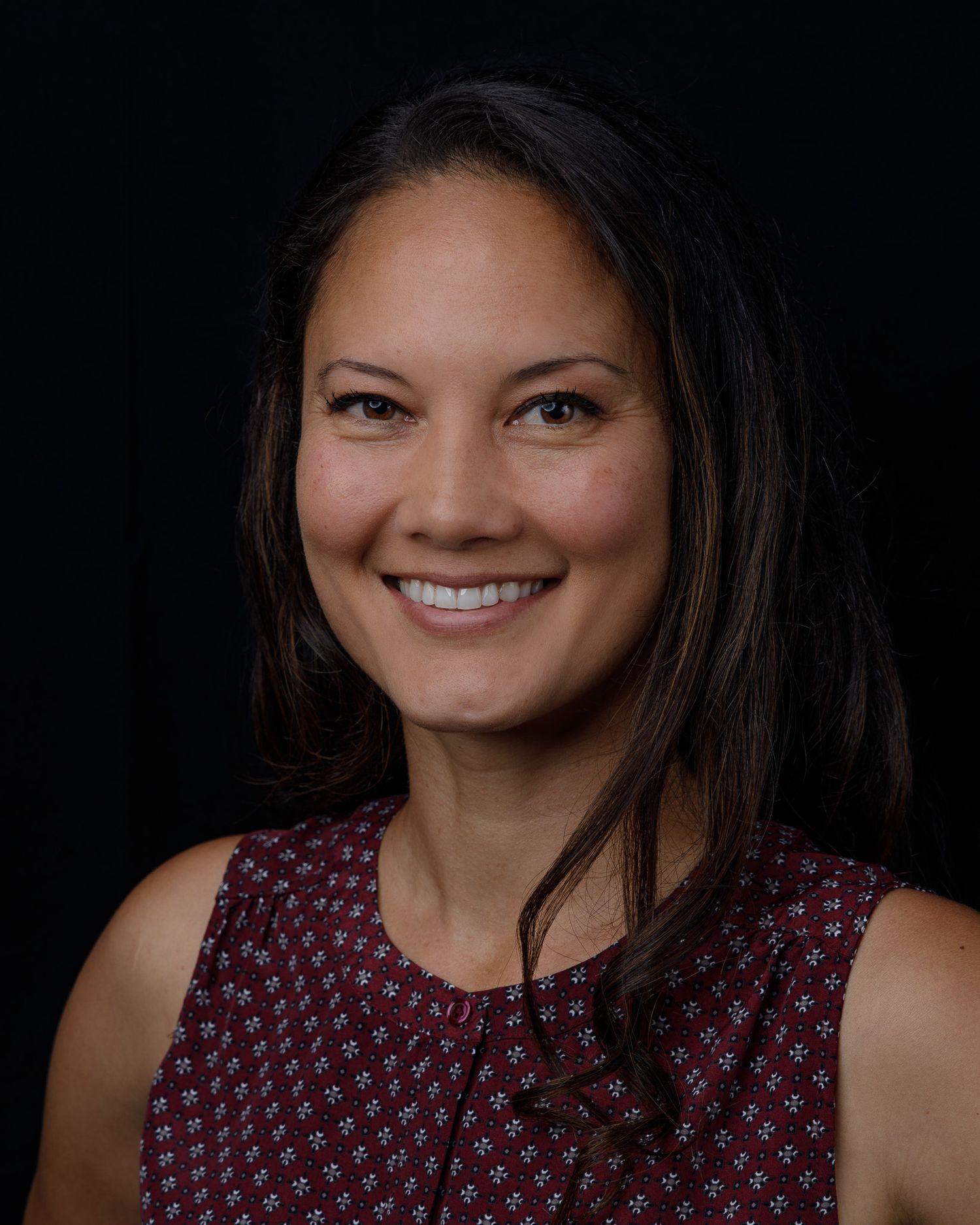Professional headshot portraits showing warm smiles against dark background wearing burgundy patterned top.
