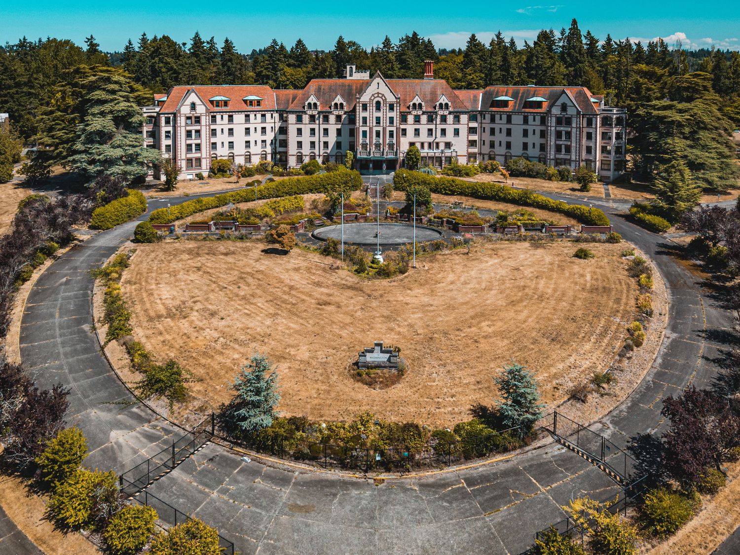 Aerial view of a large abandoned institutional building complex with circular driveway and surrounding trees.