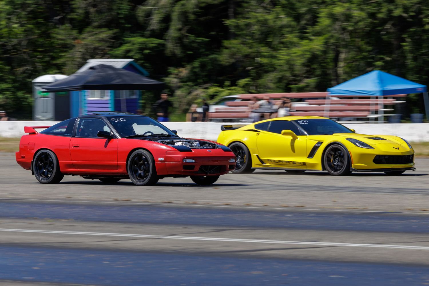 A red convertible sports car races alongside a yellow Corvette on a track surrounded by trees.