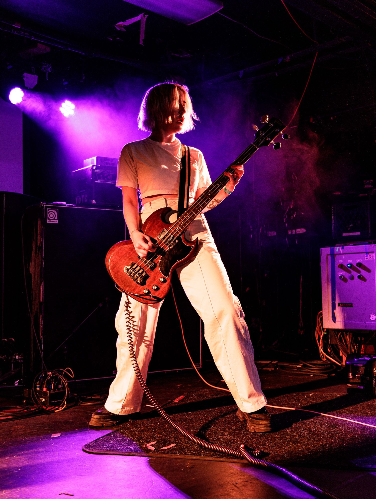 Guitarist in white pants performs energetically on stage under purple stage lighting.