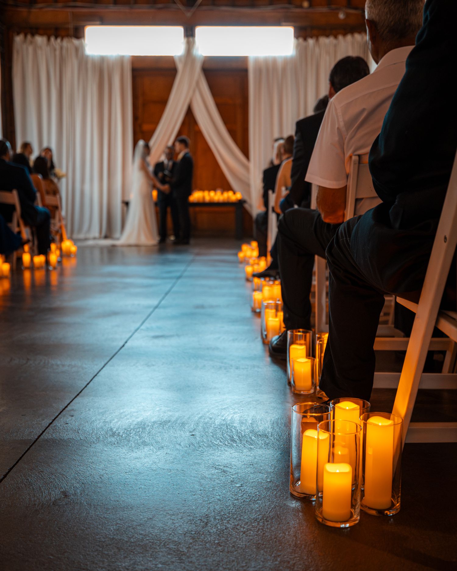 Glowing candles line a dark indoor aisle creating romantic ambiance for a wedding ceremony.