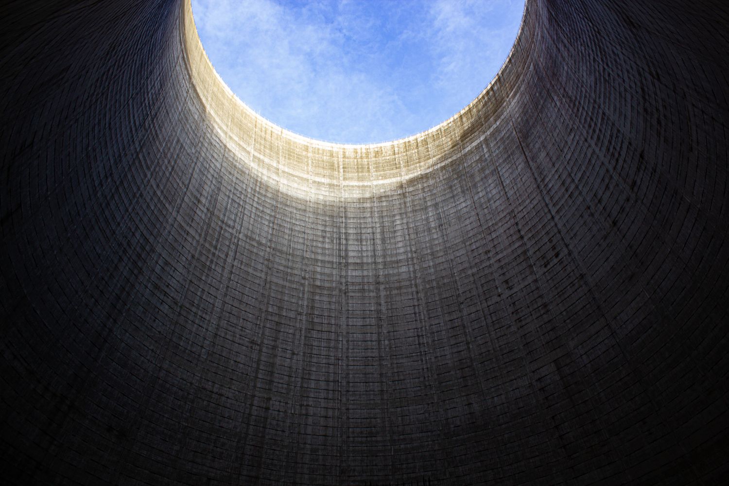 Looking up through a massive cooling tower reveals a circular opening against a bright blue sky.