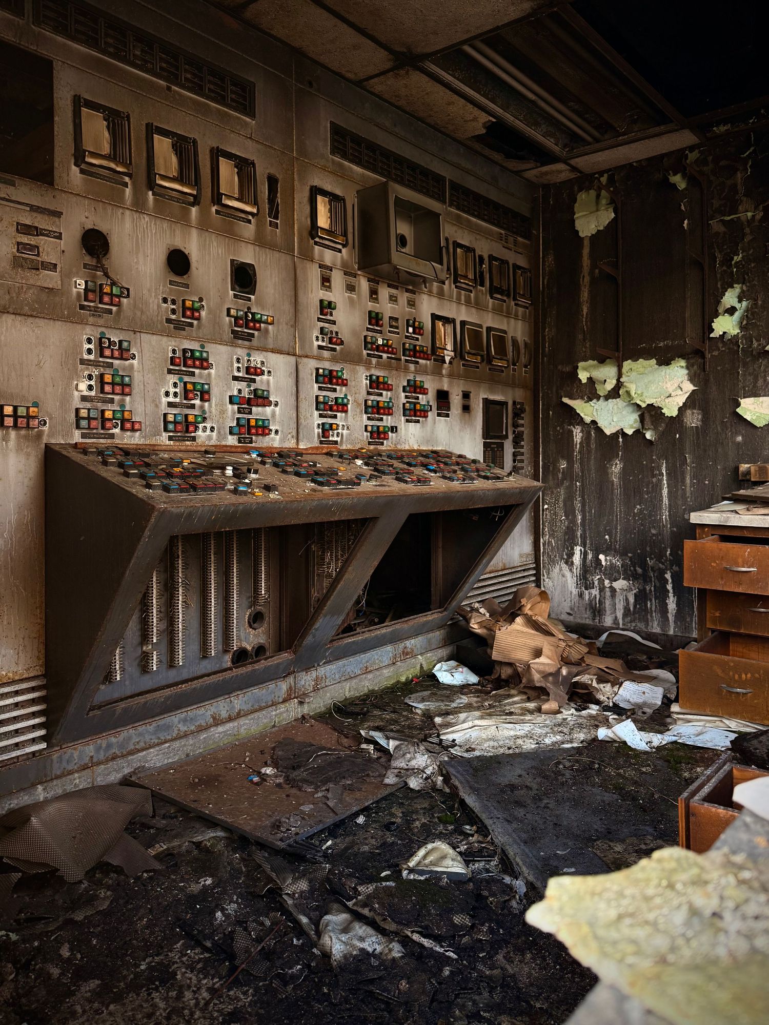 Abandoned industrial control room with decaying panels, colorful buttons, and debris-covered floor in a derelict facility.