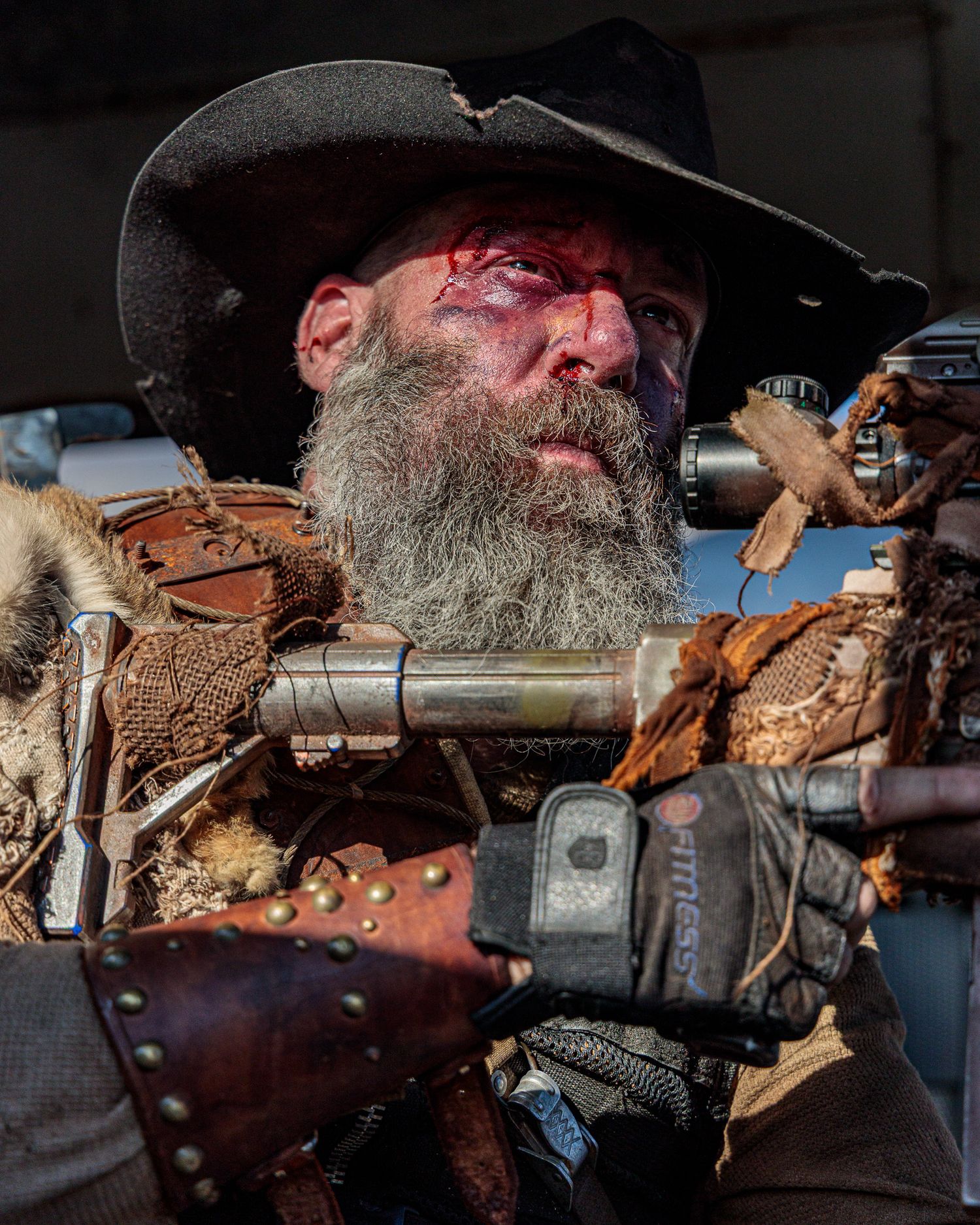 A rugged figure with weathered features wearing a cowboy hat among leather gear and equipment.