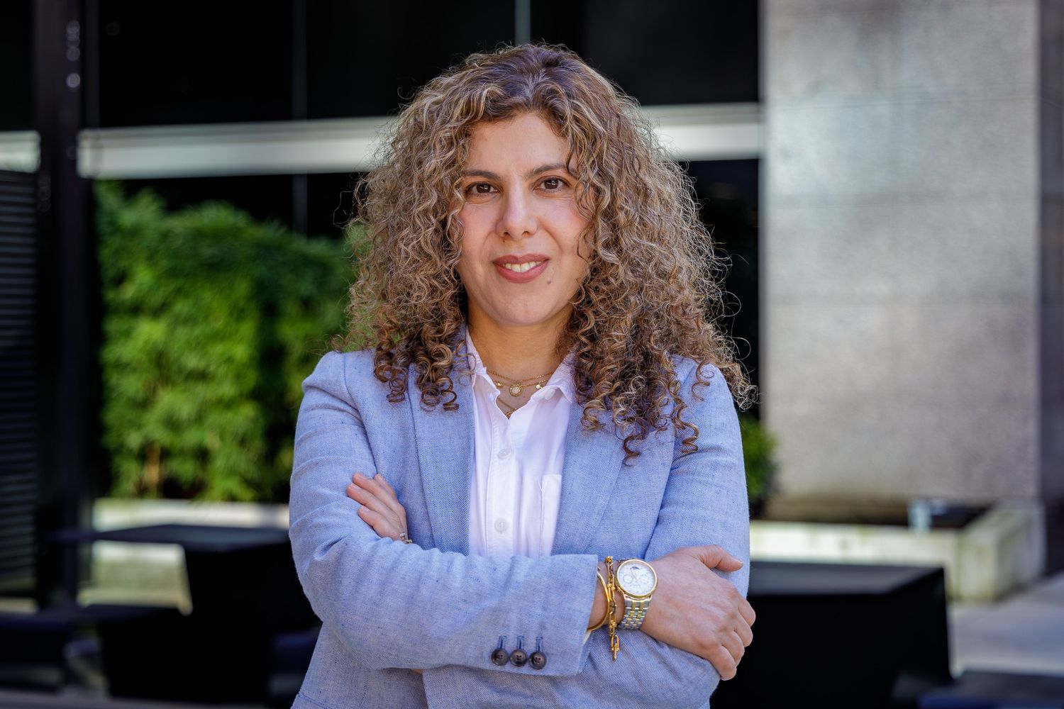 Professional headshot of a person in a light blue blazer with curly hair against an office building.