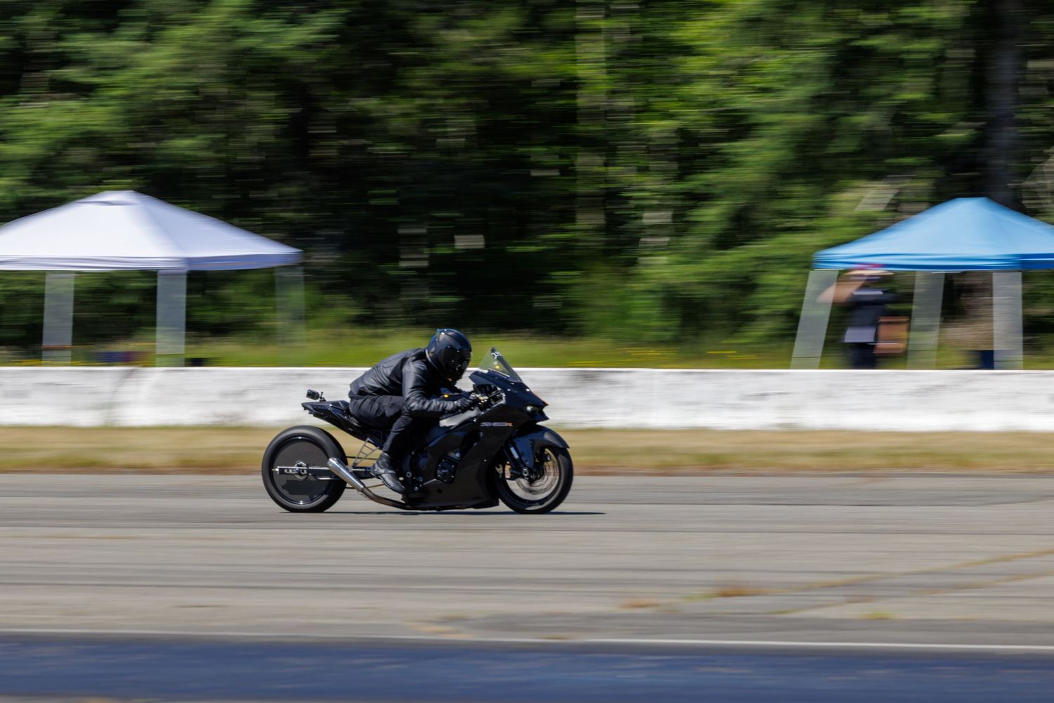 Motorcyclist in black gear speeds along a racetrack with blurred trees and blue canopy tents in the background.