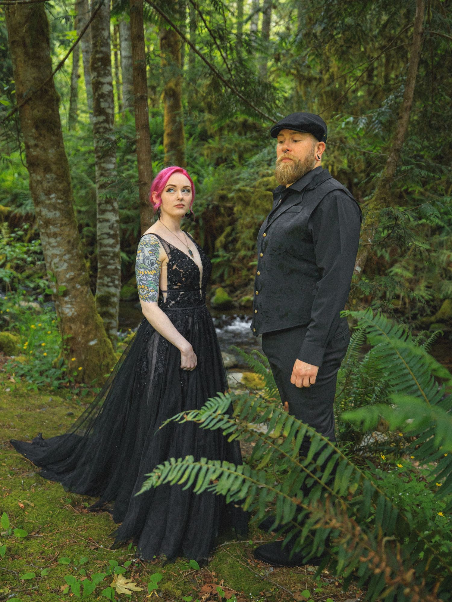 A gothic-styled couple poses in a mossy forest setting with ferns and evergreens.