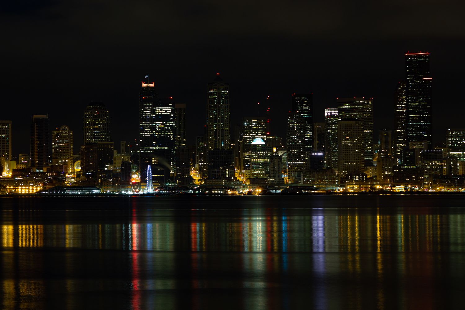Night cityscape reflecting on water with illuminated buildings and colorful lights.