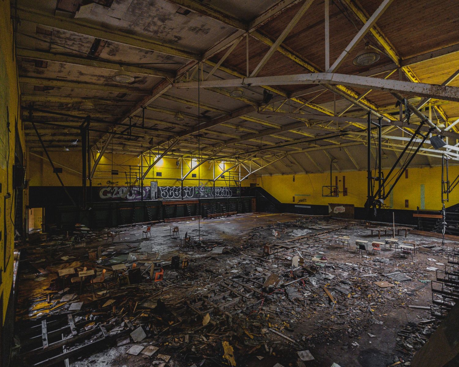 Abandoned gymnasium with yellow walls, debris-covered floor, and deteriorating roof in a derelict building.