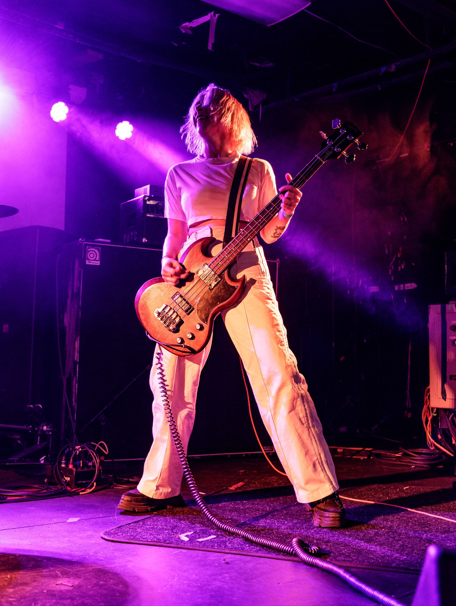 Bass player in white outfit rocks out during performance with dramatic purple stage lighting.